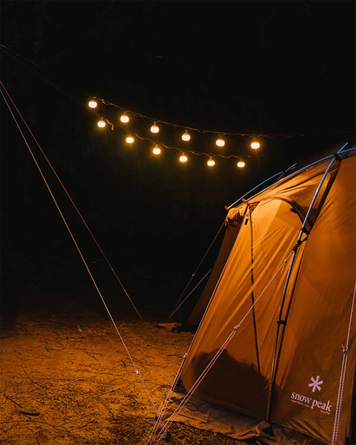 Brown Snow Peak tent illuminated by string lights at night on dirt ground.