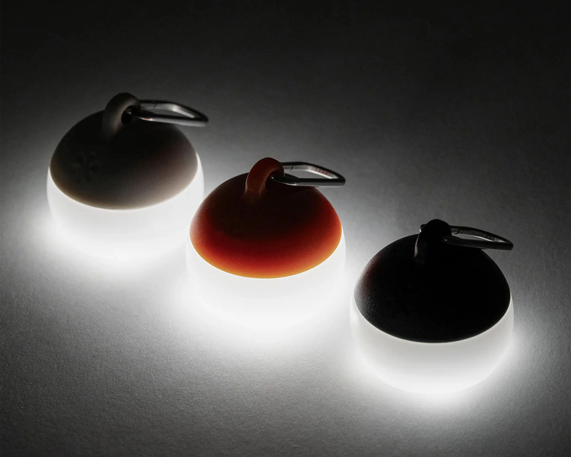 Three small round LED keychain lights with black, orange, and white tops glowing on a dark surface.