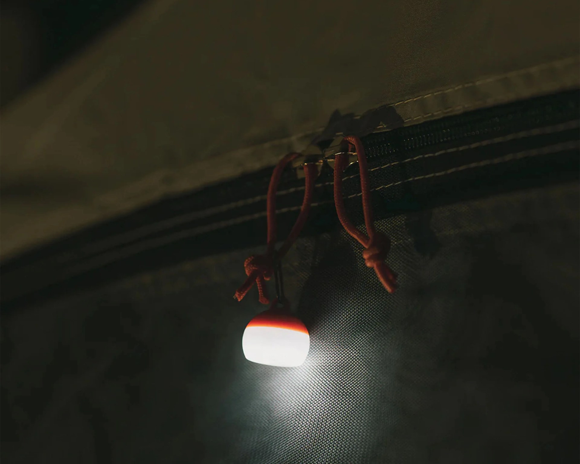 Small white and orange light hanging from a black zipper with red pull cords on dark fabric.
