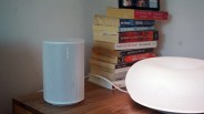 White Sonos speaker on a wooden surface next to a stack of books and a glowing white lamp.