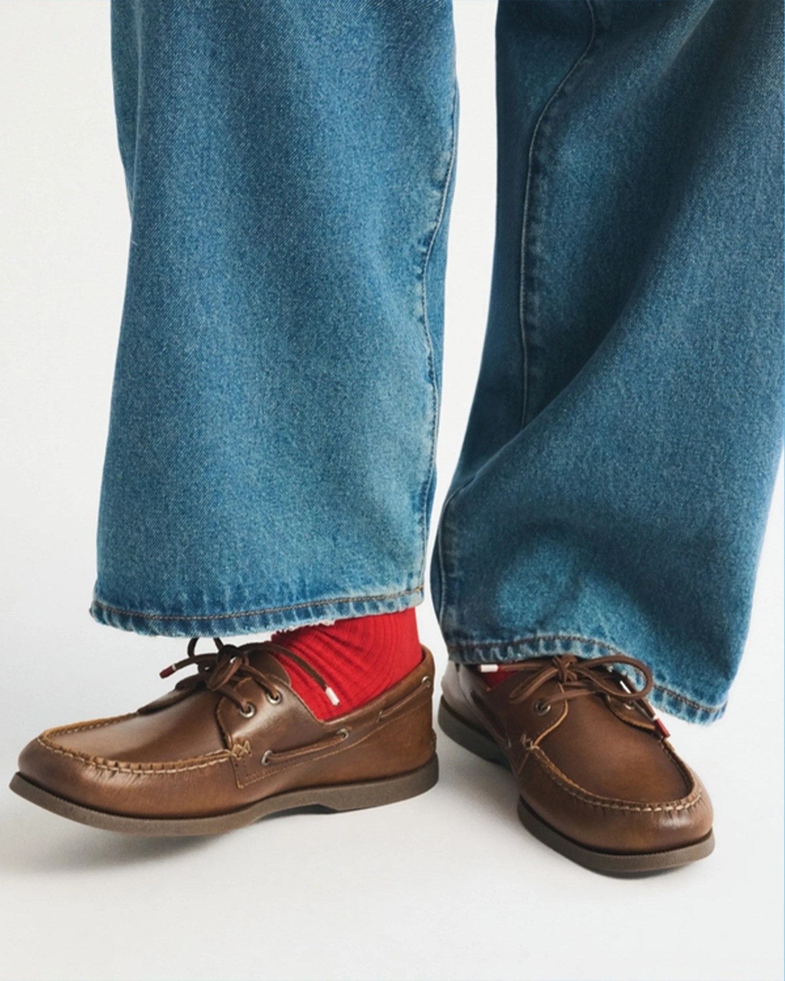 Brown leather boat shoes worn with red socks and blue denim jeans.