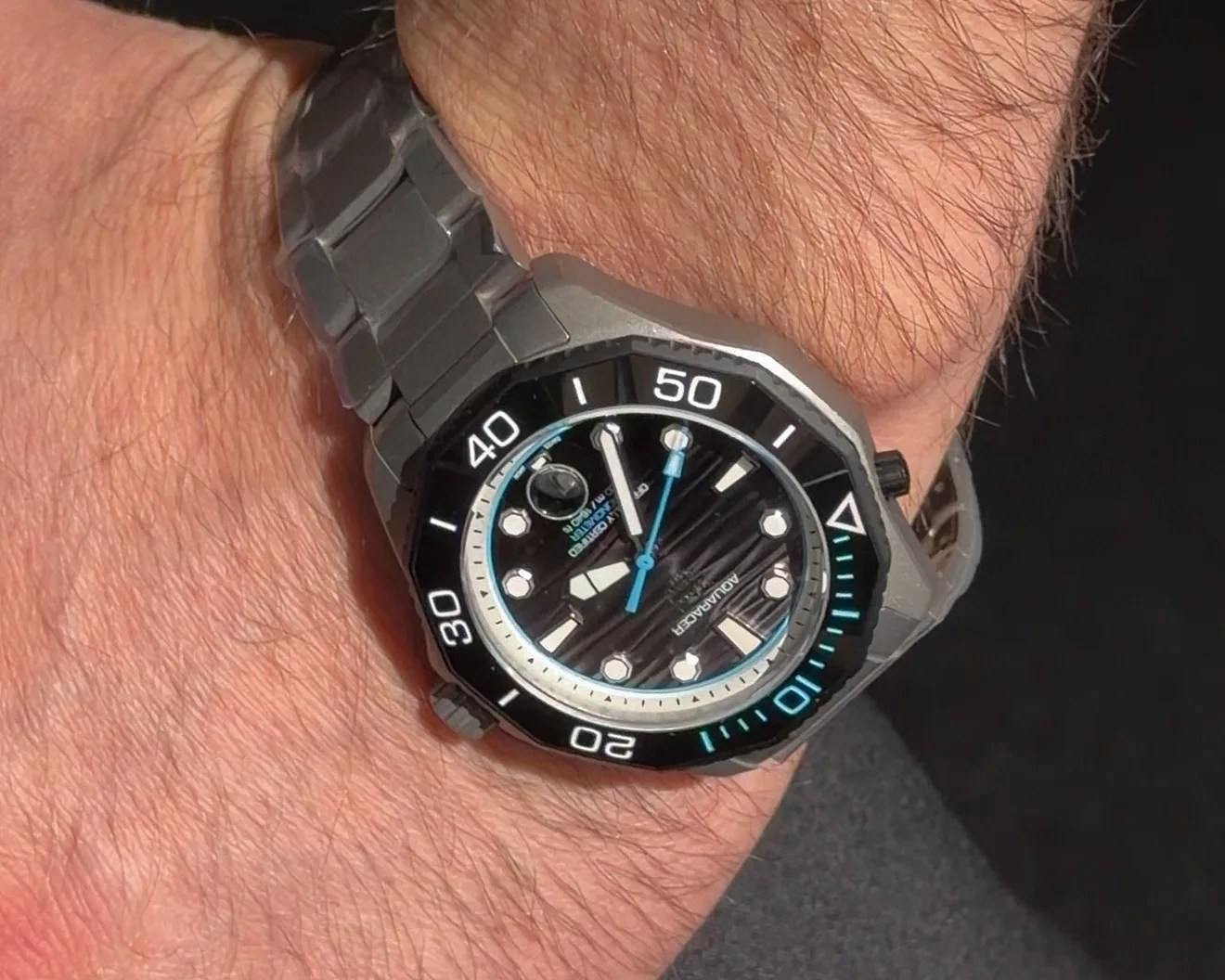 Black dial dive watch with white and blue markers and a gray metal bracelet on a wrist.