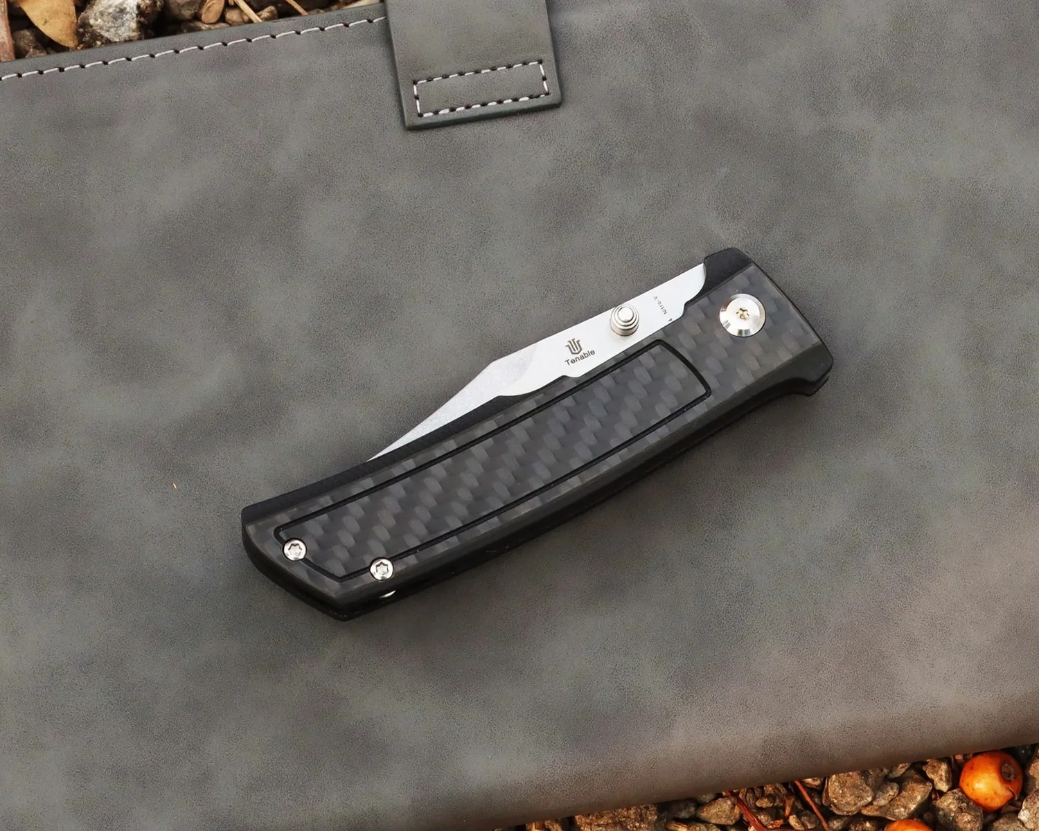 Folded pocket knife with carbon fiber handle and partially visible stainless steel blade on a gray leather surface.