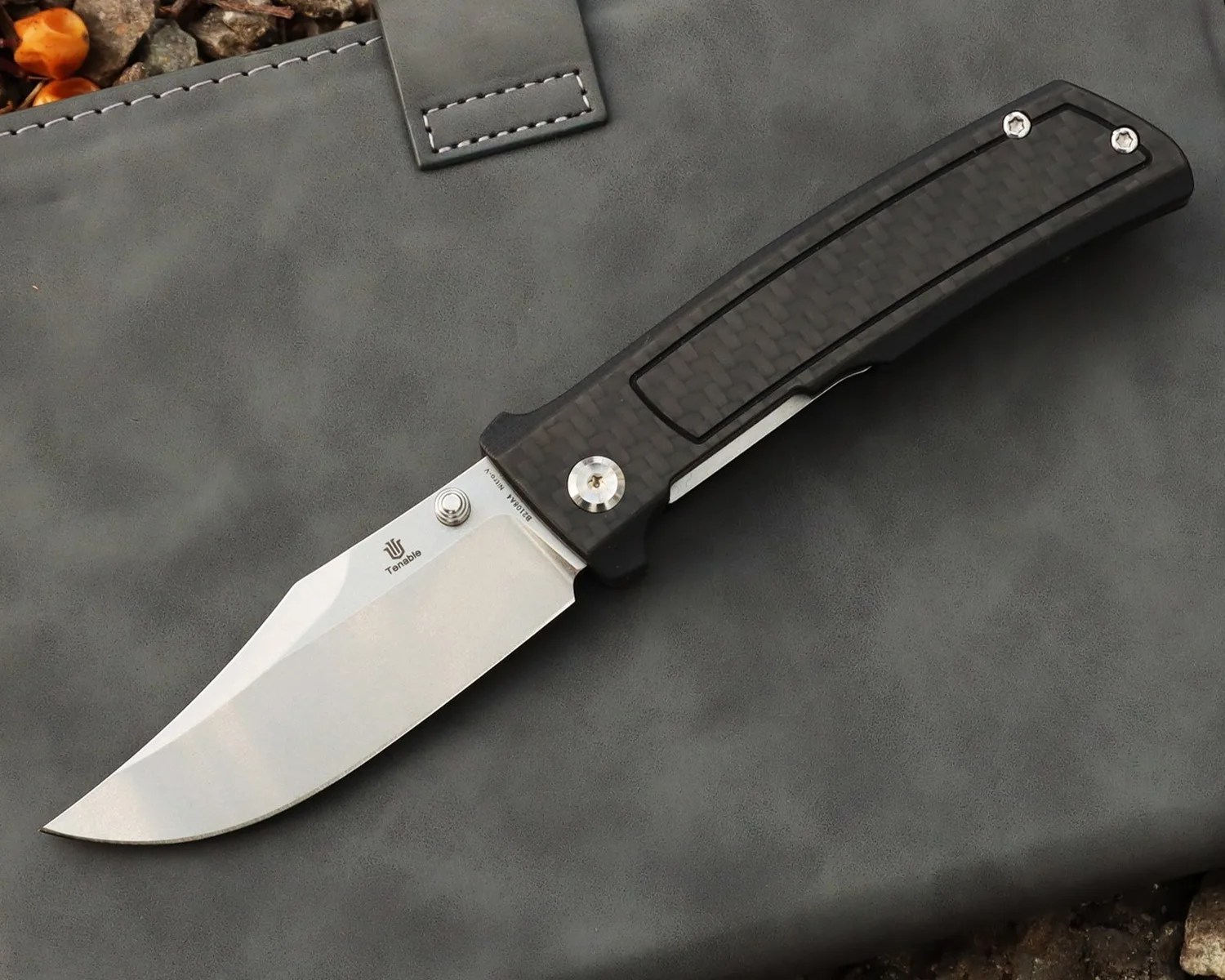 Folding knife with a silver blade and black carbon fiber handle resting on a gray leather surface.