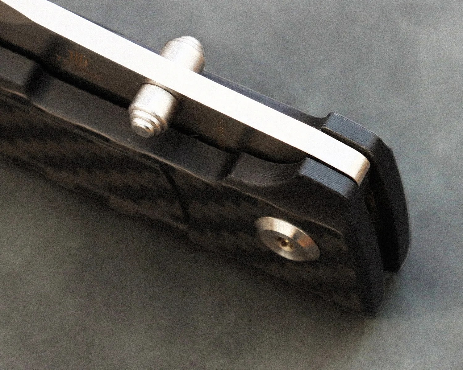 Close-up of a folding knife's pivot and handle showing a carbon fiber texture and metal hardware.