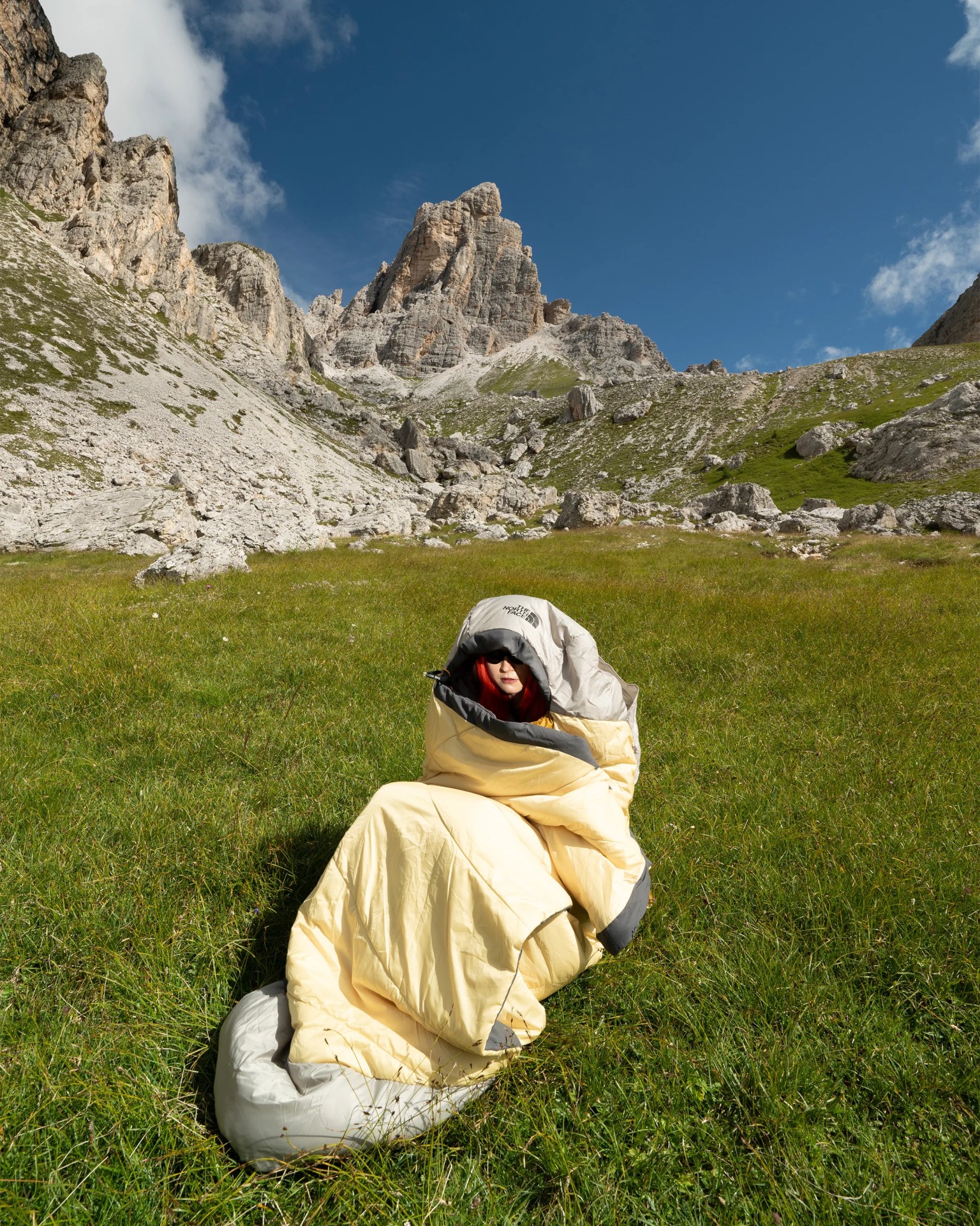 Person wrapped in a yellow and gray sleeping bag sitting on green grass with rocky mountains in the background.