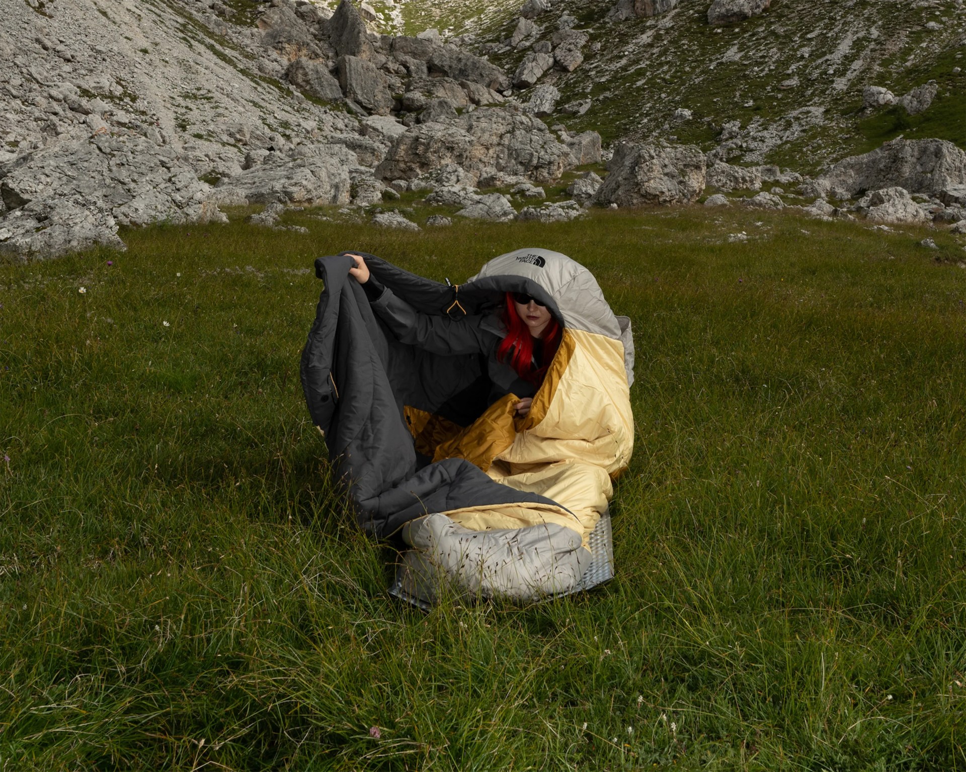 Person with red hair wrapped in a gray and yellow North Face sleeping bag sitting on grass with rocky hills in the background.