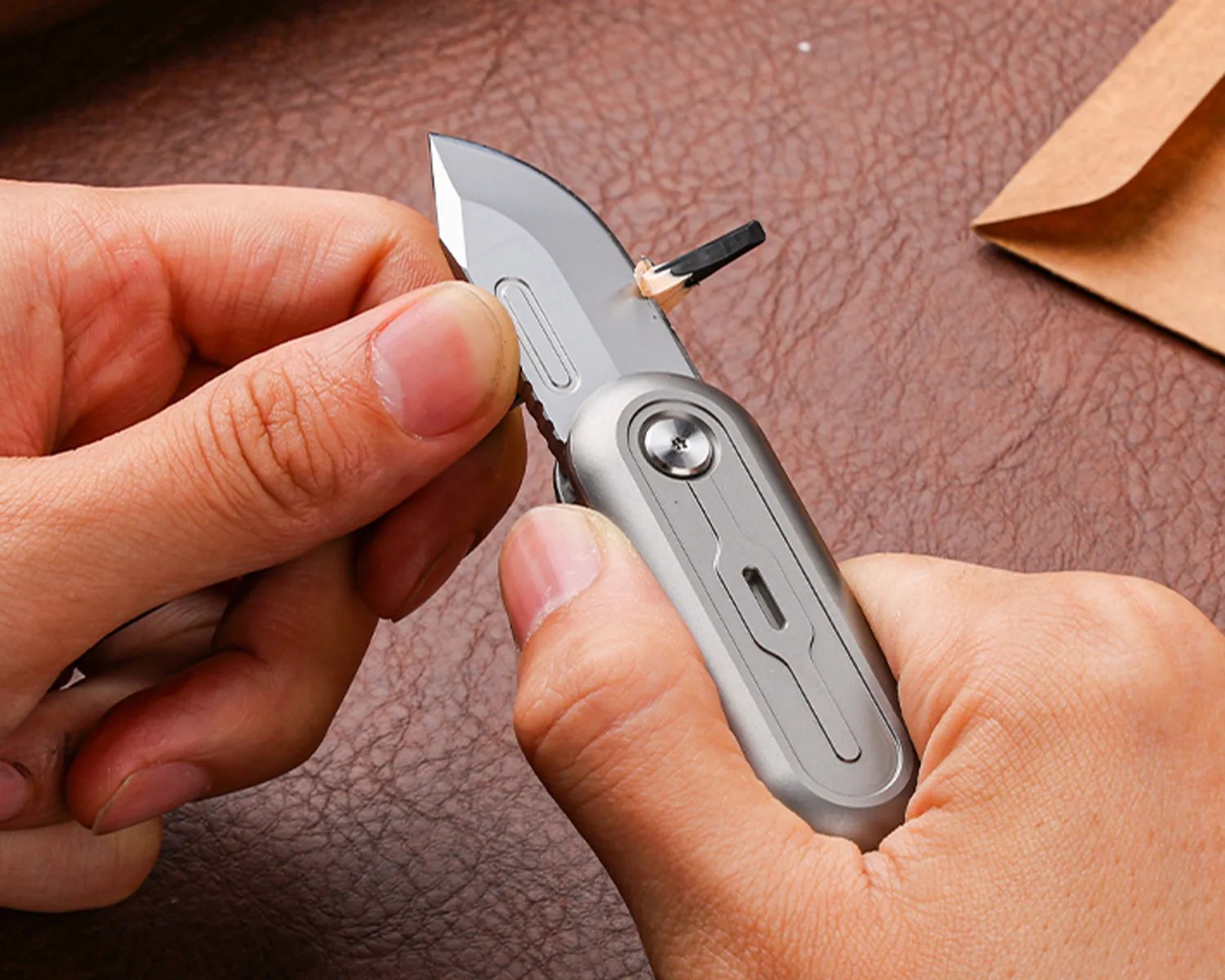 Hands sharpening a black pencil tip using a silver utility knife on a brown textured surface.