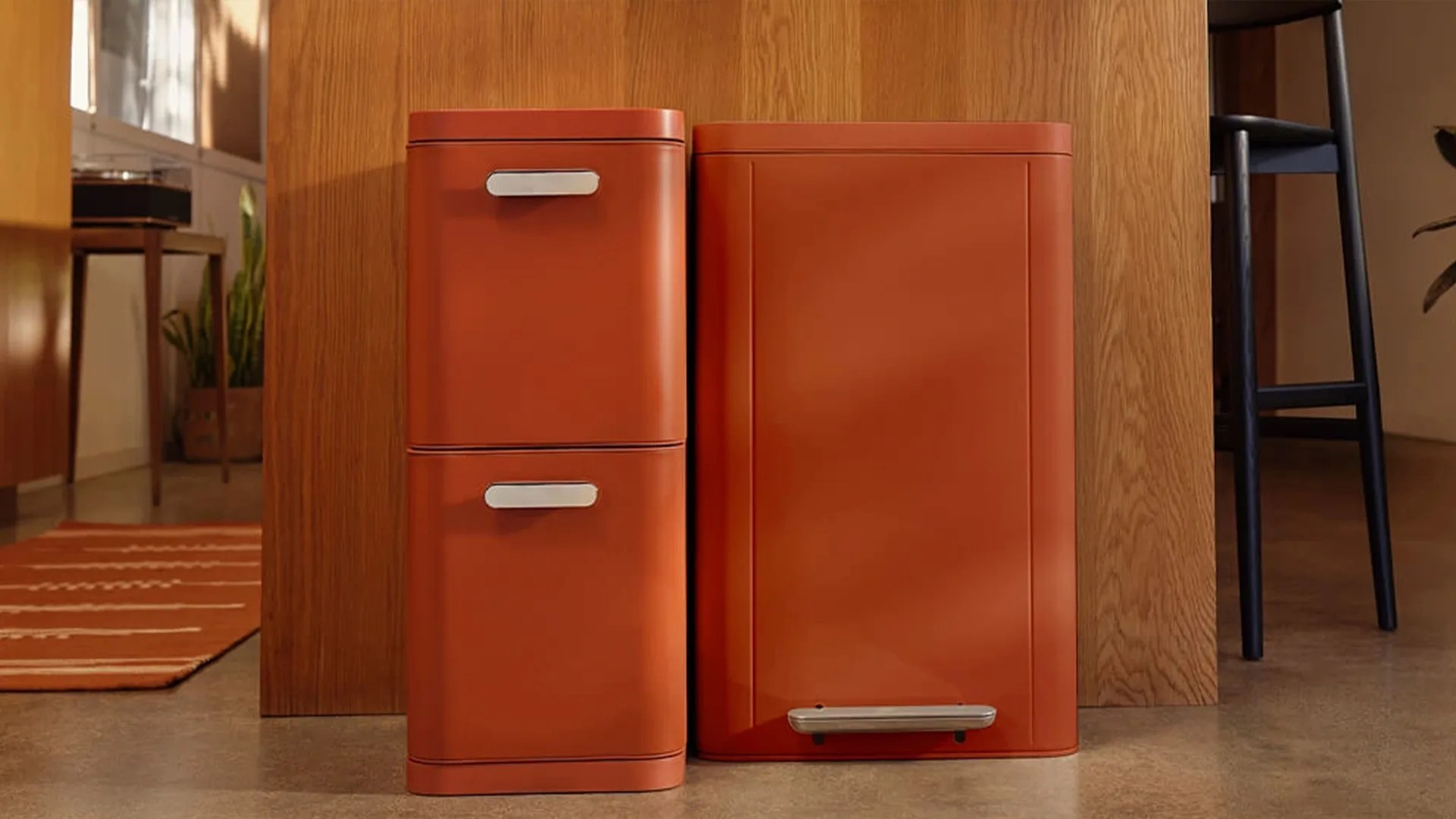 Two rust-orange metal trash bins with silver handles and pedal, placed on a floor against a wooden panel.