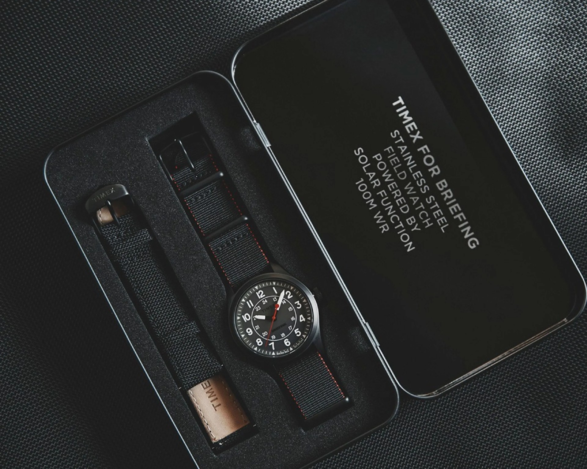 Black Timex field watch with white numerals and red second hand in a black foam-lined metal box with an extra black strap.