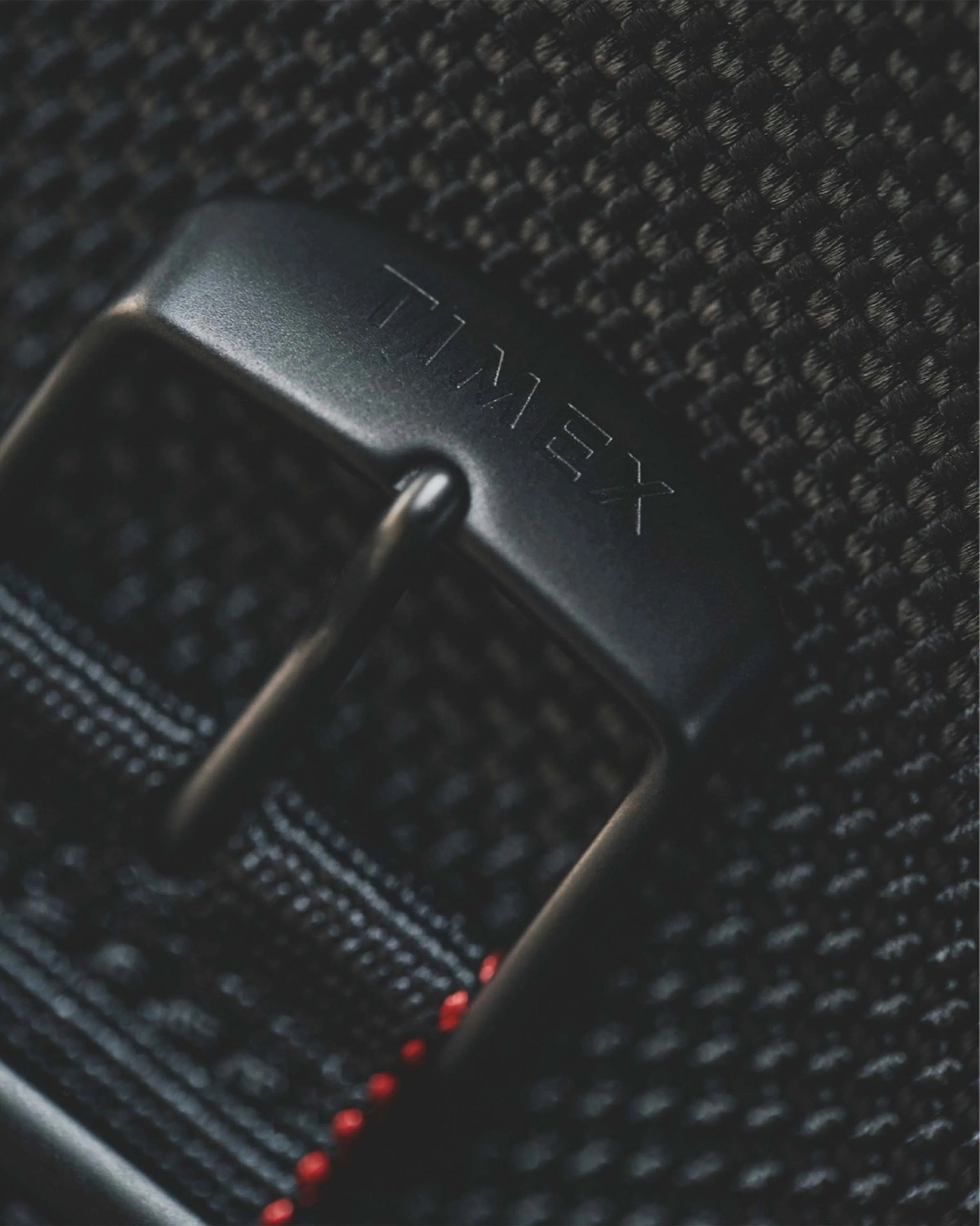 Close-up of a black Timex buckle on a textured black fabric strap with red stitching.