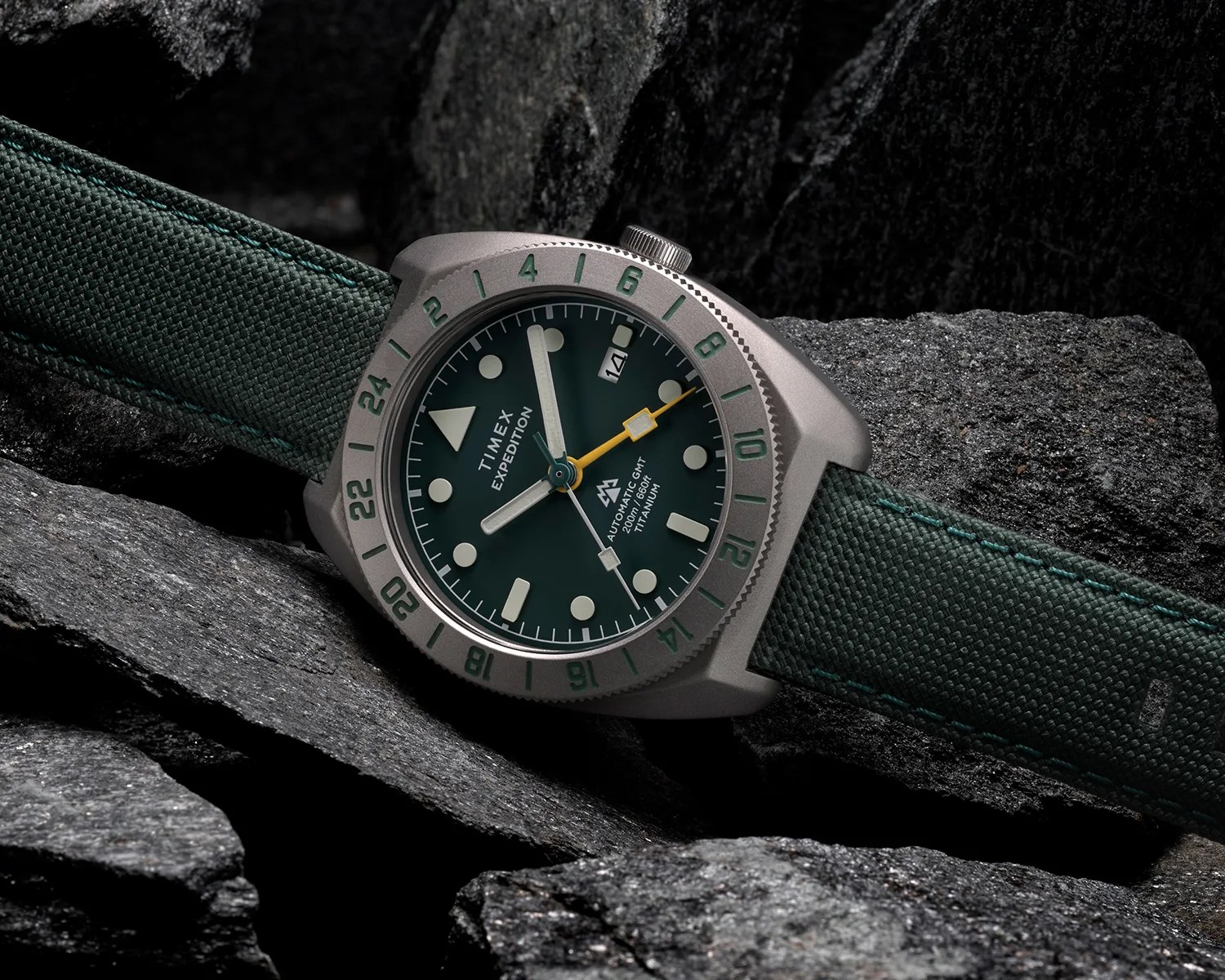 Timex Expedition watch with green dial, yellow GMT hand, titanium case, and green fabric strap on dark rocks.