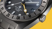 Close-up of a titanium automatic GMT watch with a black dial, yellow GMT hand, and black numbered bezel on a yellow background.