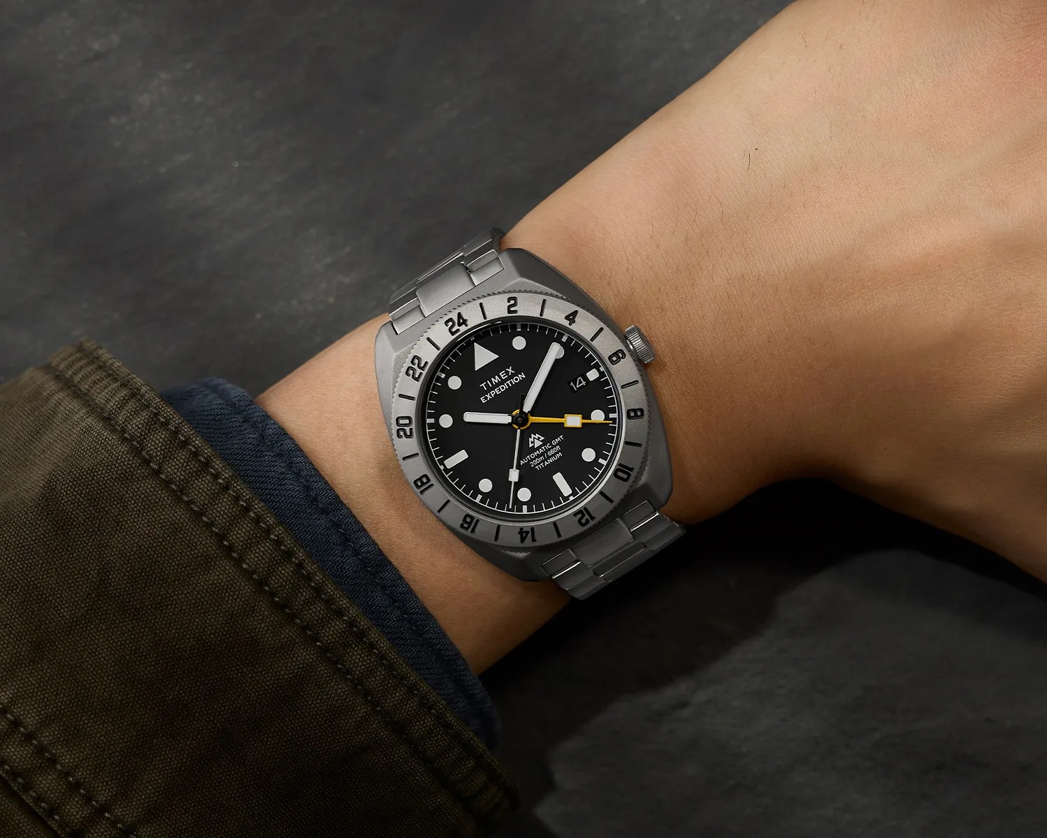 Titanium Timex Expedition wristwatch with black dial, white markers, yellow GMT hand, and metal bracelet on a wrist.