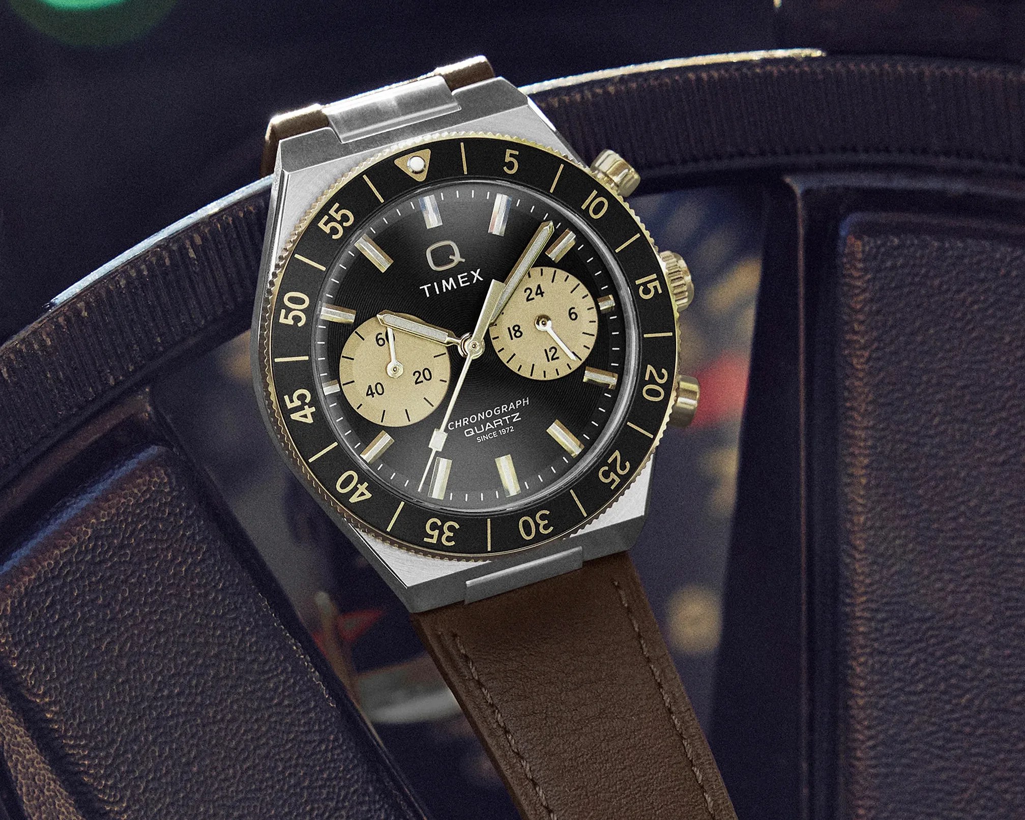 Timex Q chronograph quartz watch with black dial, gold-tone bezel, and brown leather strap on a dark surface.