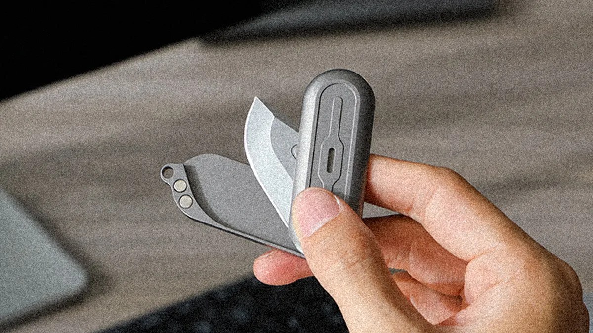 Hand holding a small gray multi-tool with a folding blade and additional tools on a wooden surface.