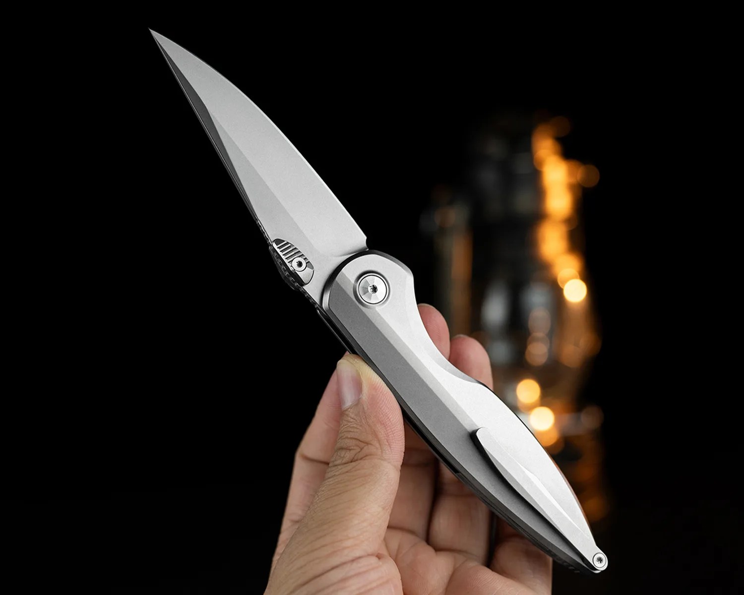 Hand holding a sleek silver folding knife with a sharp pointed blade against a dark background with blurred warm lights.