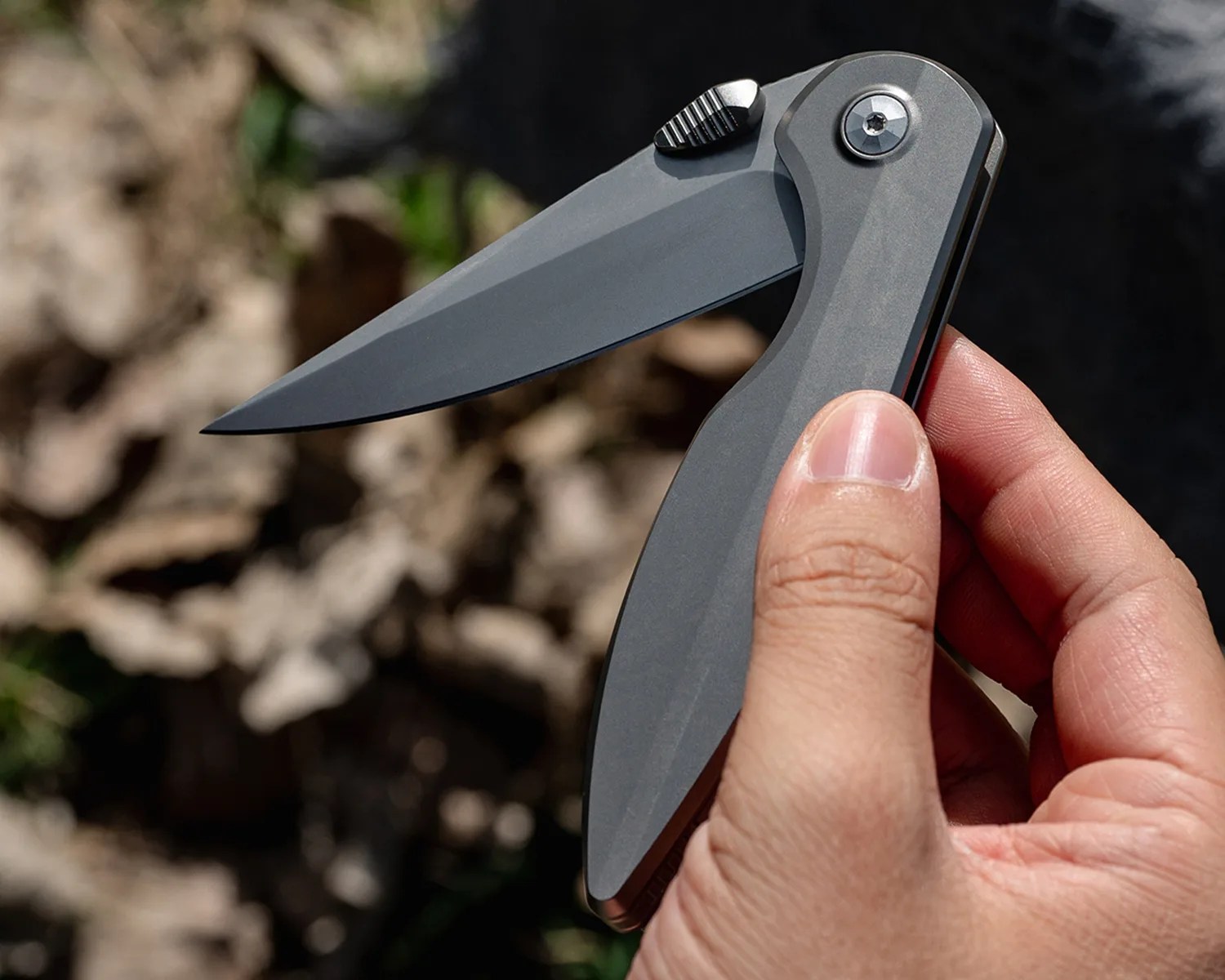 Hand holding a sleek gray folding knife with a partially open blade outdoors.