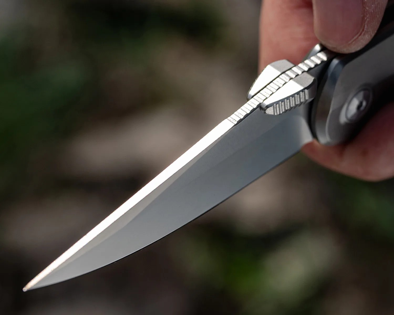 Close-up of a sharp, silver folding knife blade with textured thumb grip held in a hand.