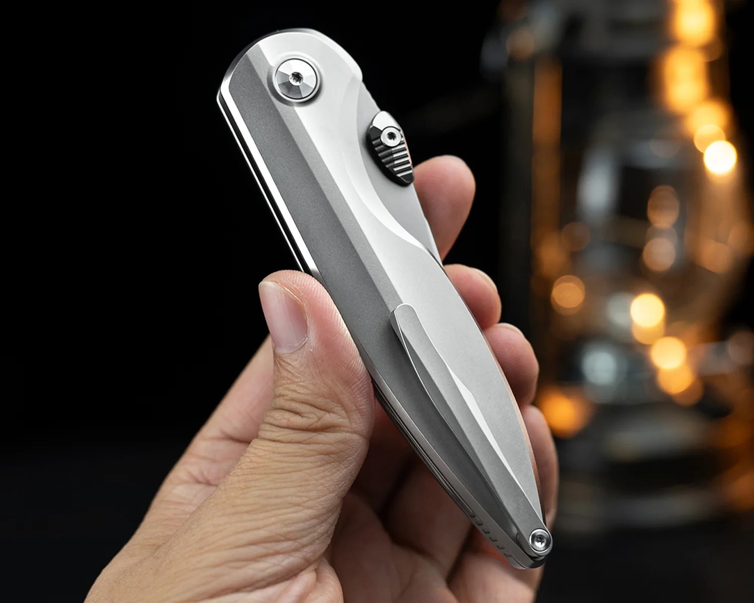 Hand holding a closed silver folding knife with a clip and textured thumb stud.