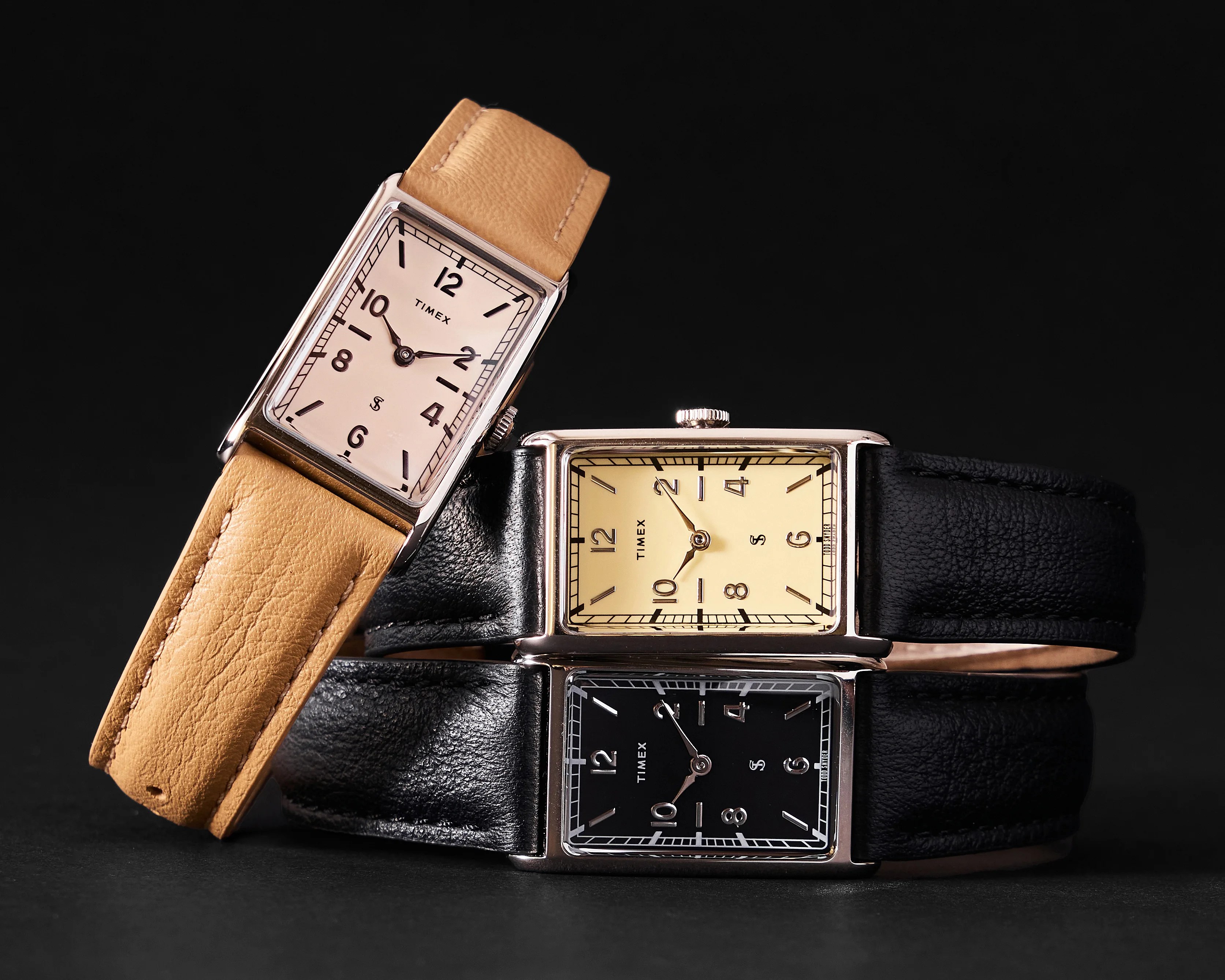 Three rectangular Timex wristwatches with leather straps in tan and black, stacked on a black reflective surface.