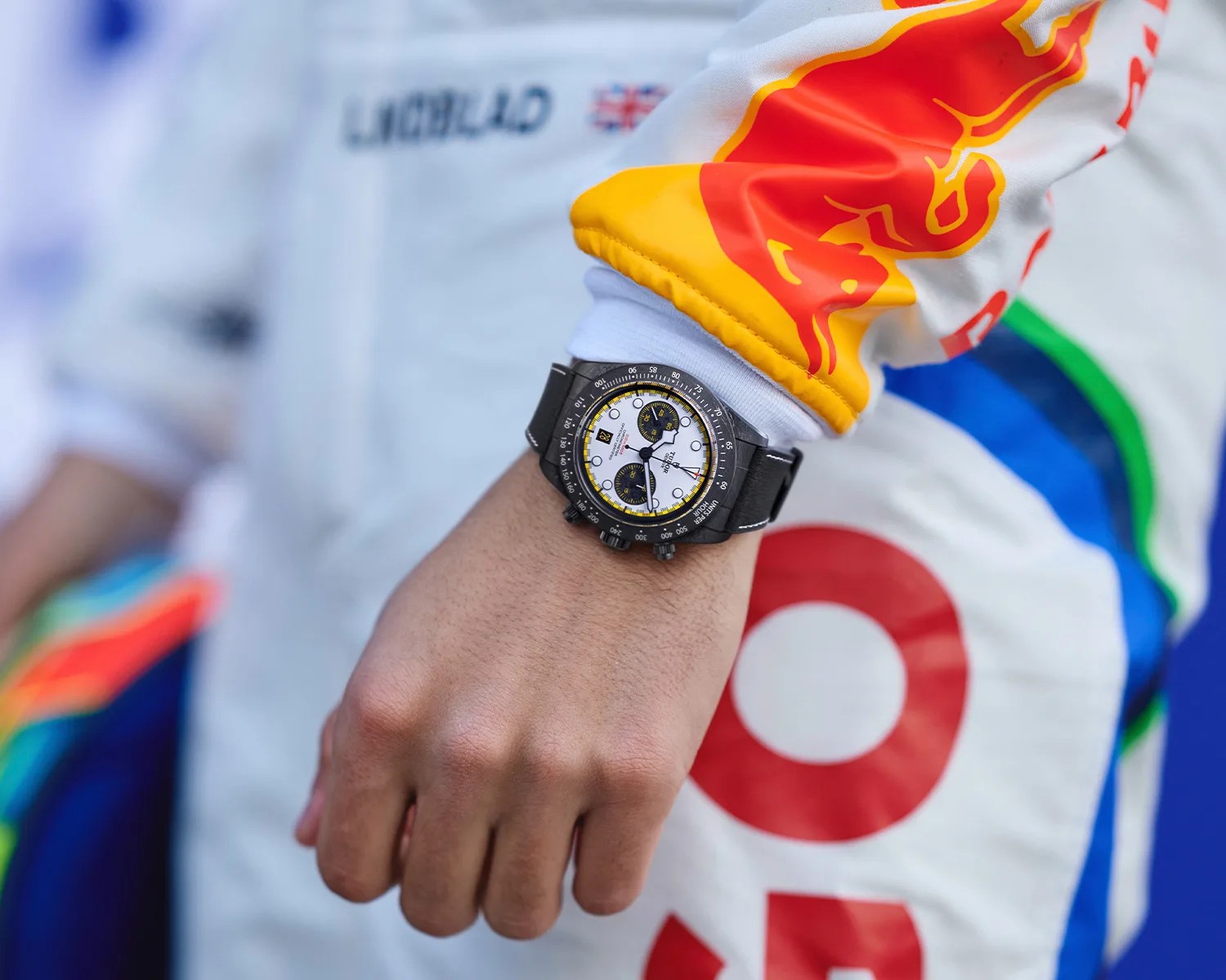 Black chronograph wristwatch with white dial and yellow accents worn on a wrist over a racing suit sleeve with Red Bull logo.
