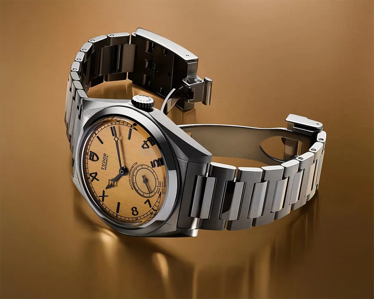 Stainless steel Tudor Monarch wristwatch with a textured golden dial and black numerals on a reflective brown surface.