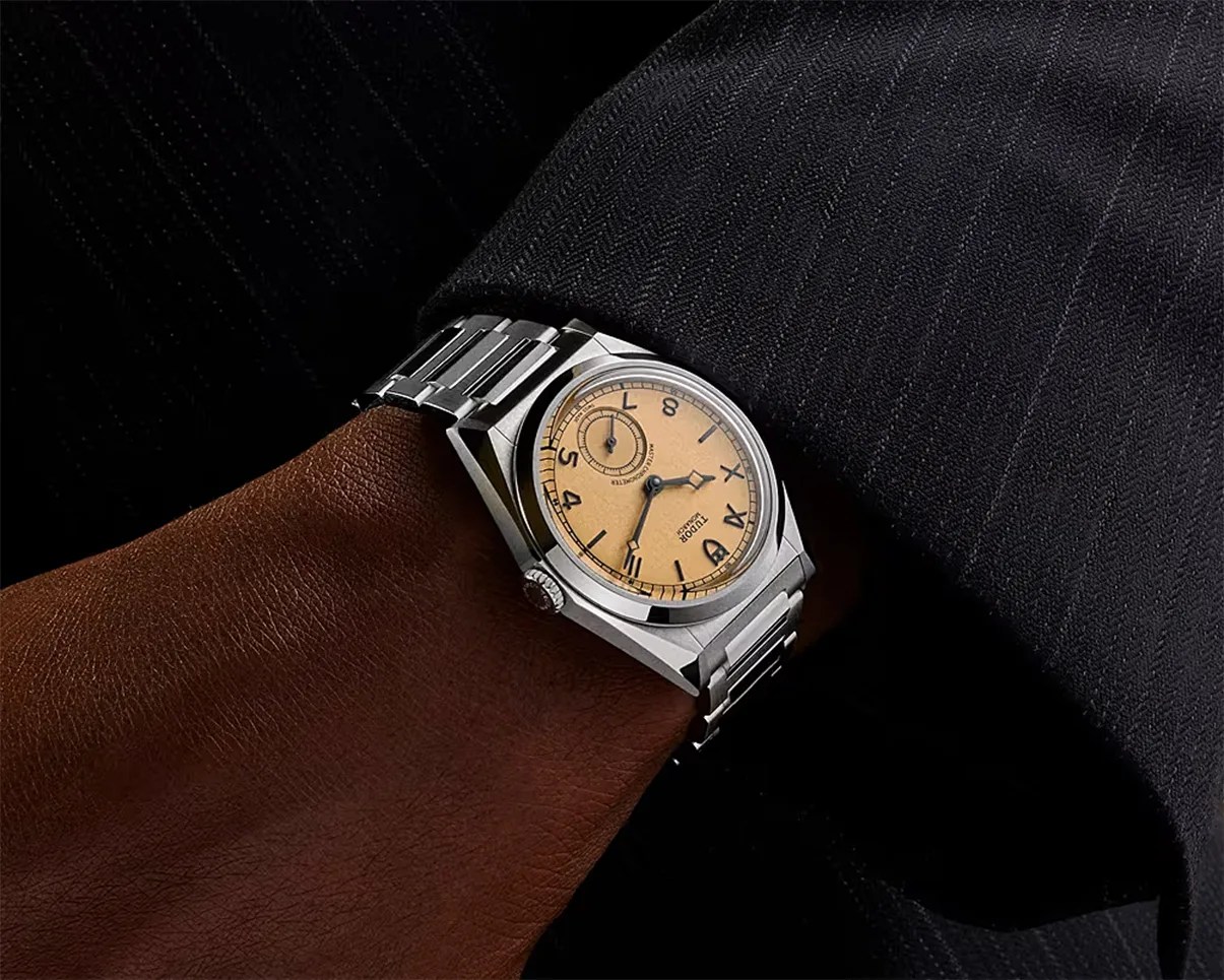 Silver Tudor wristwatch with a beige dial and black numerals worn on a wrist with a dark pinstripe sleeve.