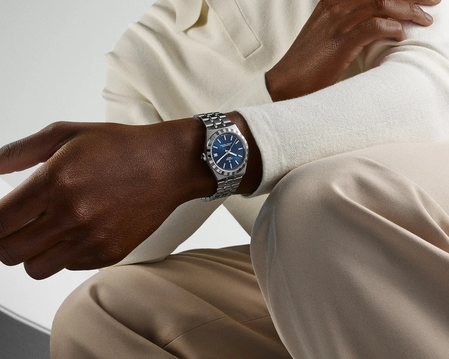 Silver metal wristwatch with a blue dial worn on a wrist over a cream long-sleeve shirt and beige pants.