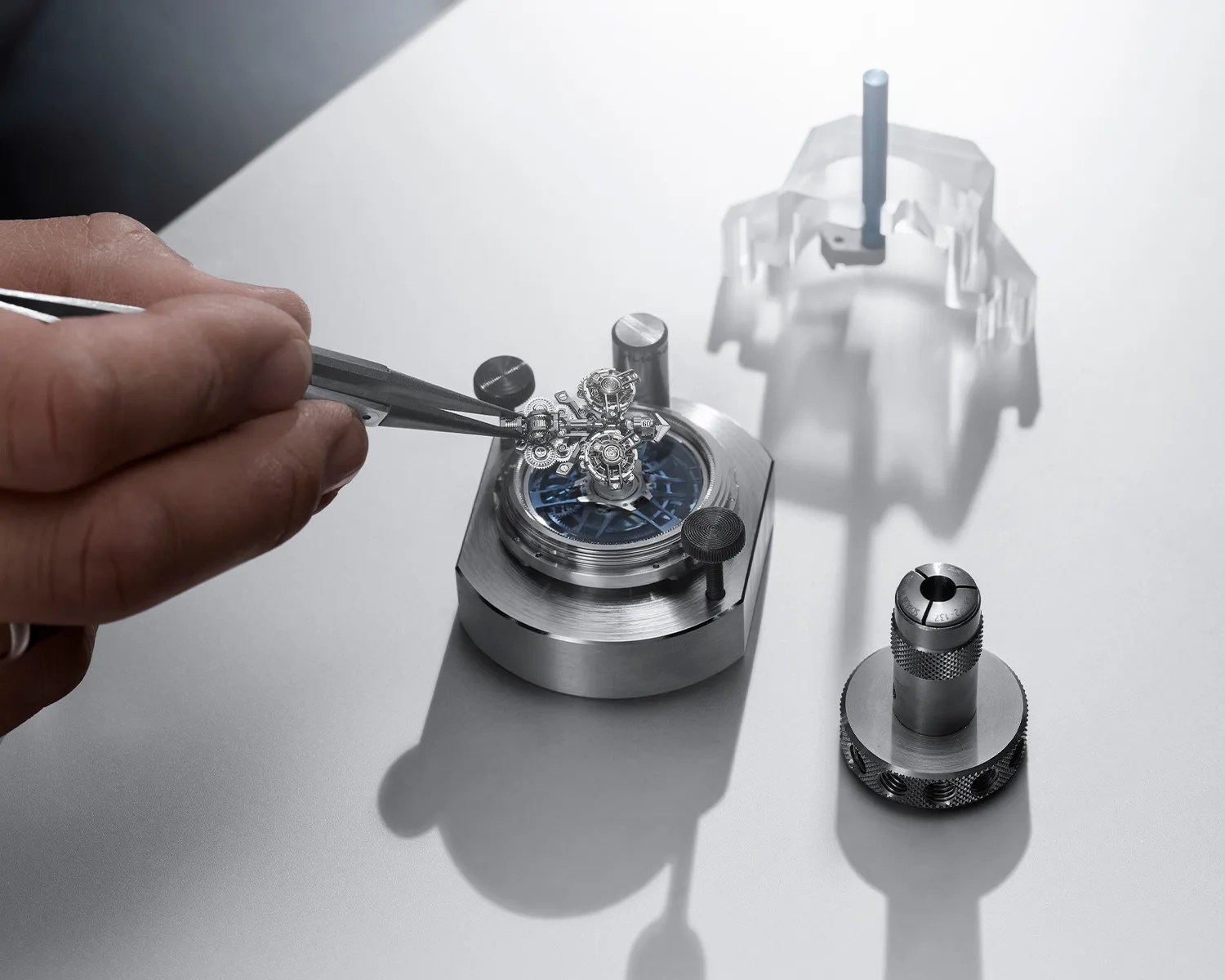 Hand holding tweezers assembling a small intricate watch mechanism on a metal base with tools on a gray surface.