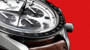 Close-up of a Universal Genève Compax chronograph watch with a white dial, black subdials, black bezel, and brown leather strap on a red background.
