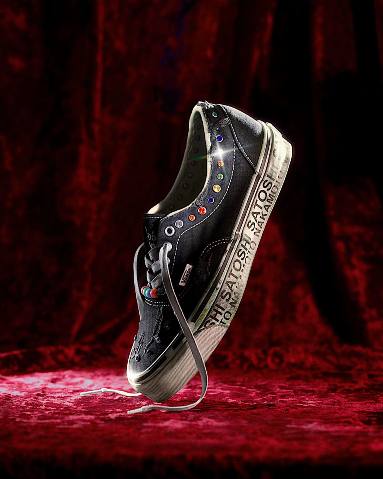 Black Vans sneaker with colorful rhinestones on eyelets and text on white sole, suspended above red velvet surface.