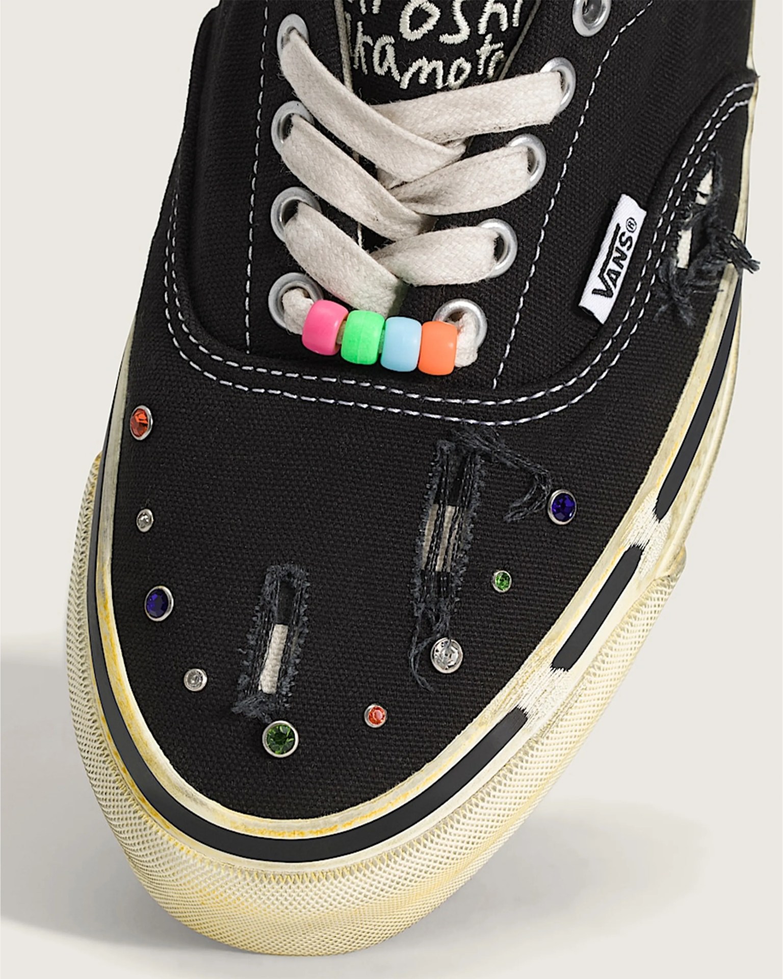 Close-up of a black Vans sneaker with white laces, colorful beads on the laces, distressed fabric, and multicolored rhinestone embellishments.