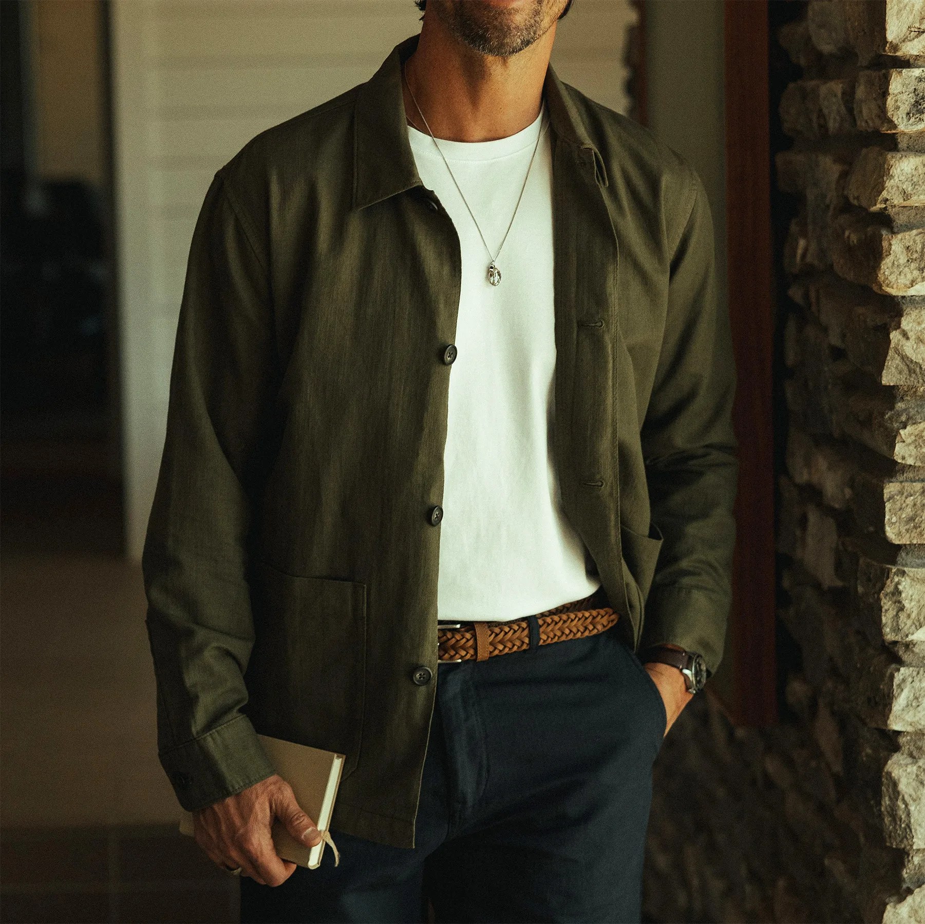 Man wearing an olive green button-up jacket over a white t-shirt, navy pants, and a brown woven belt, holding a notebook.