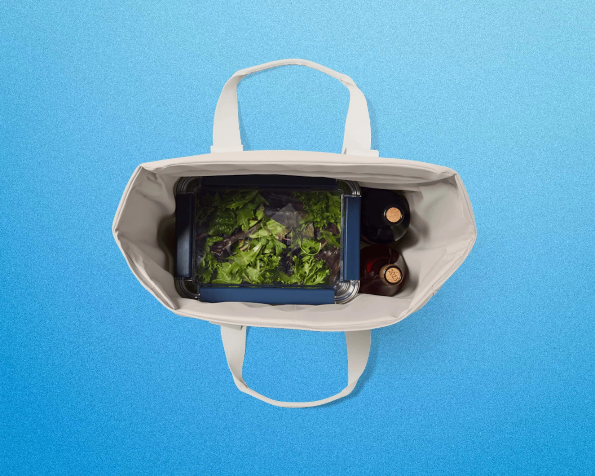 White reusable tote bag containing a plastic container of leafy greens and two glass bottles with cork stoppers.