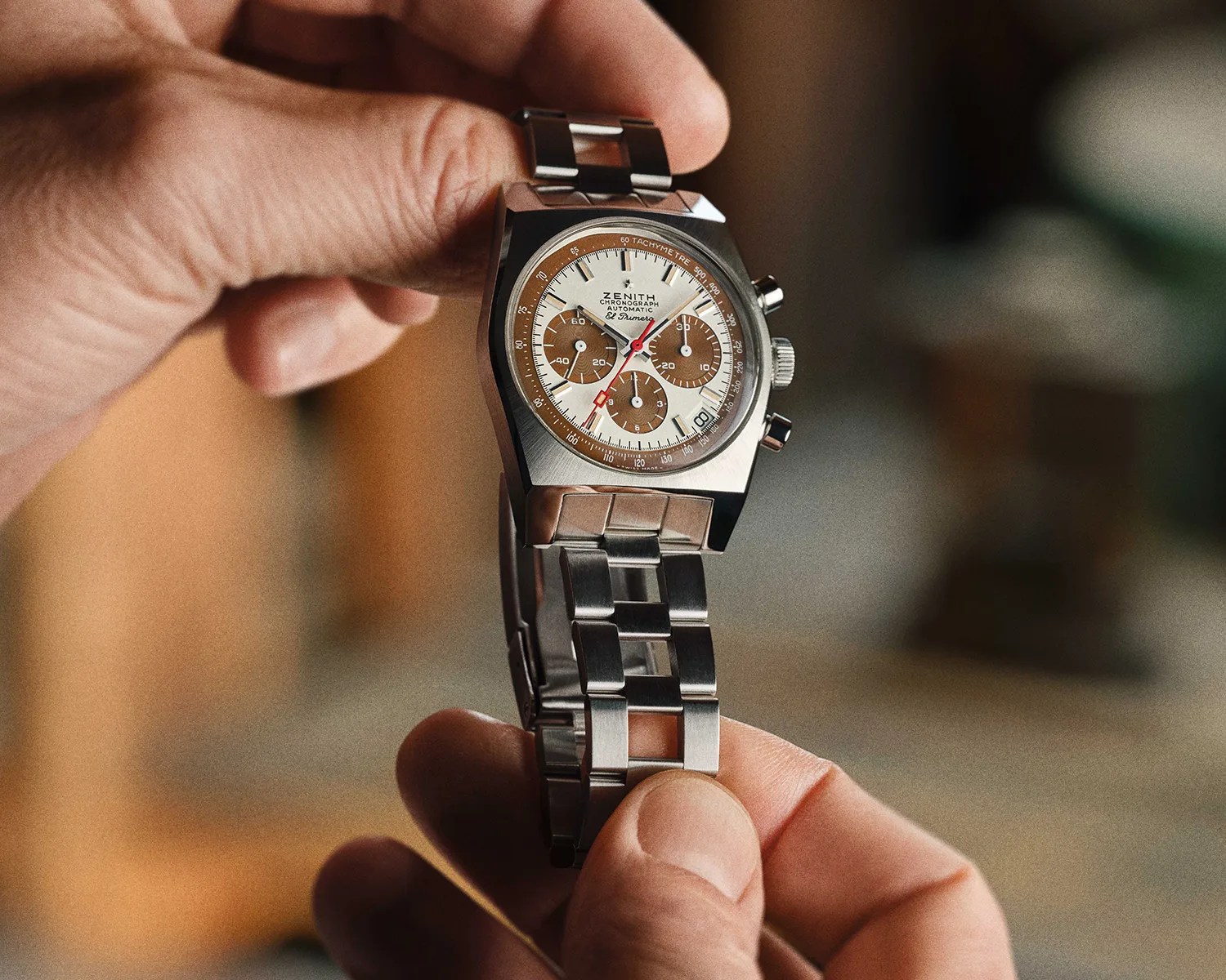 Silver Zenith El Primero chronograph wristwatch with brown subdials and tachymeter bezel held between two hands.
