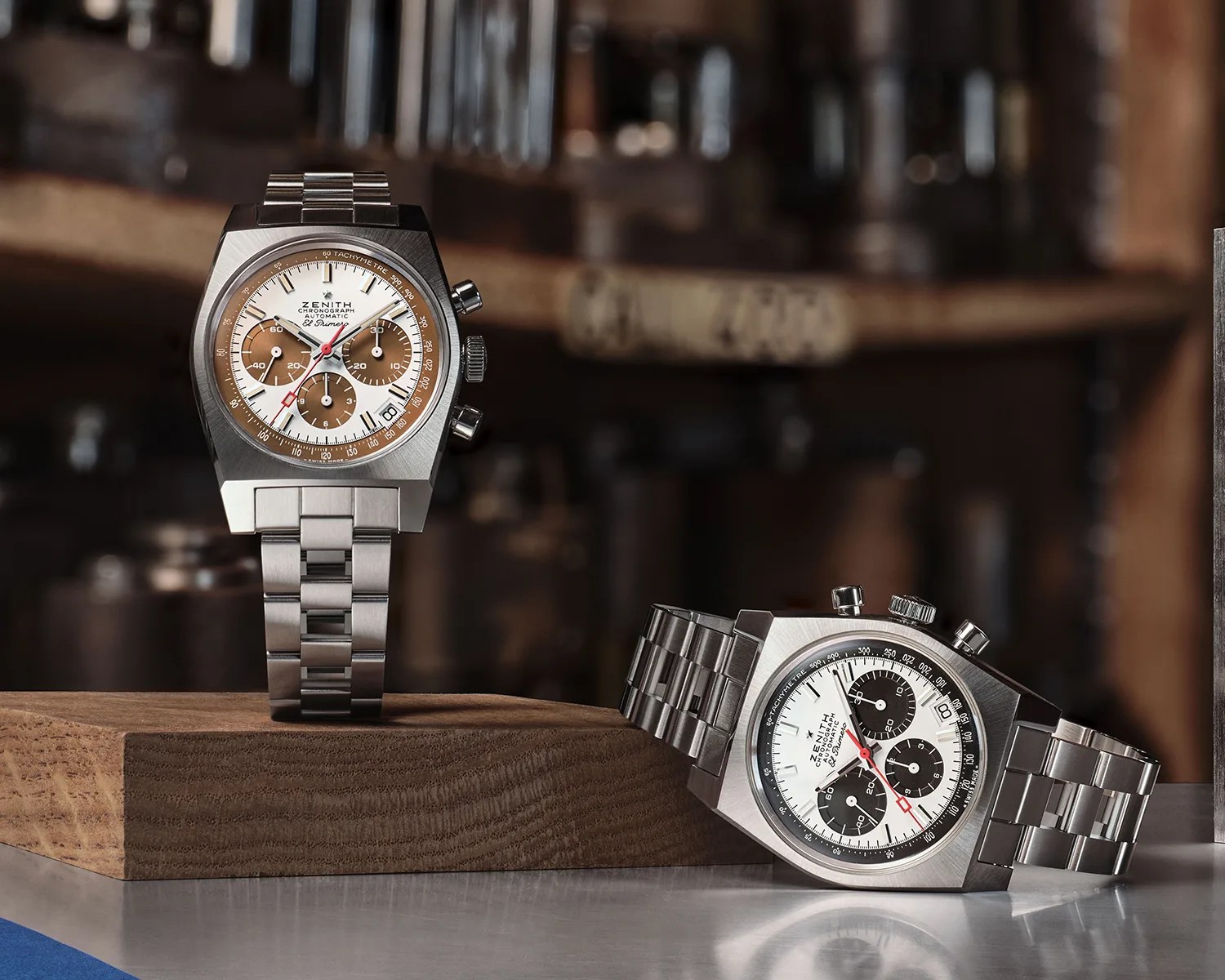 Two Zenith El Primero chronograph watches with stainless steel bracelets, one with a white and brown dial standing on a wooden block, the other with a white and black dial lying on a reflective surface.