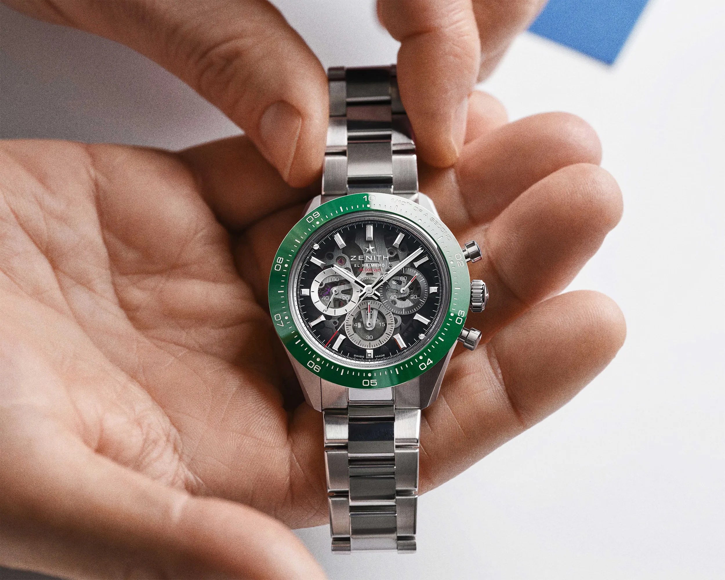 Silver Zenith El Primero chronograph watch with green bezel and metal bracelet held in a hand.