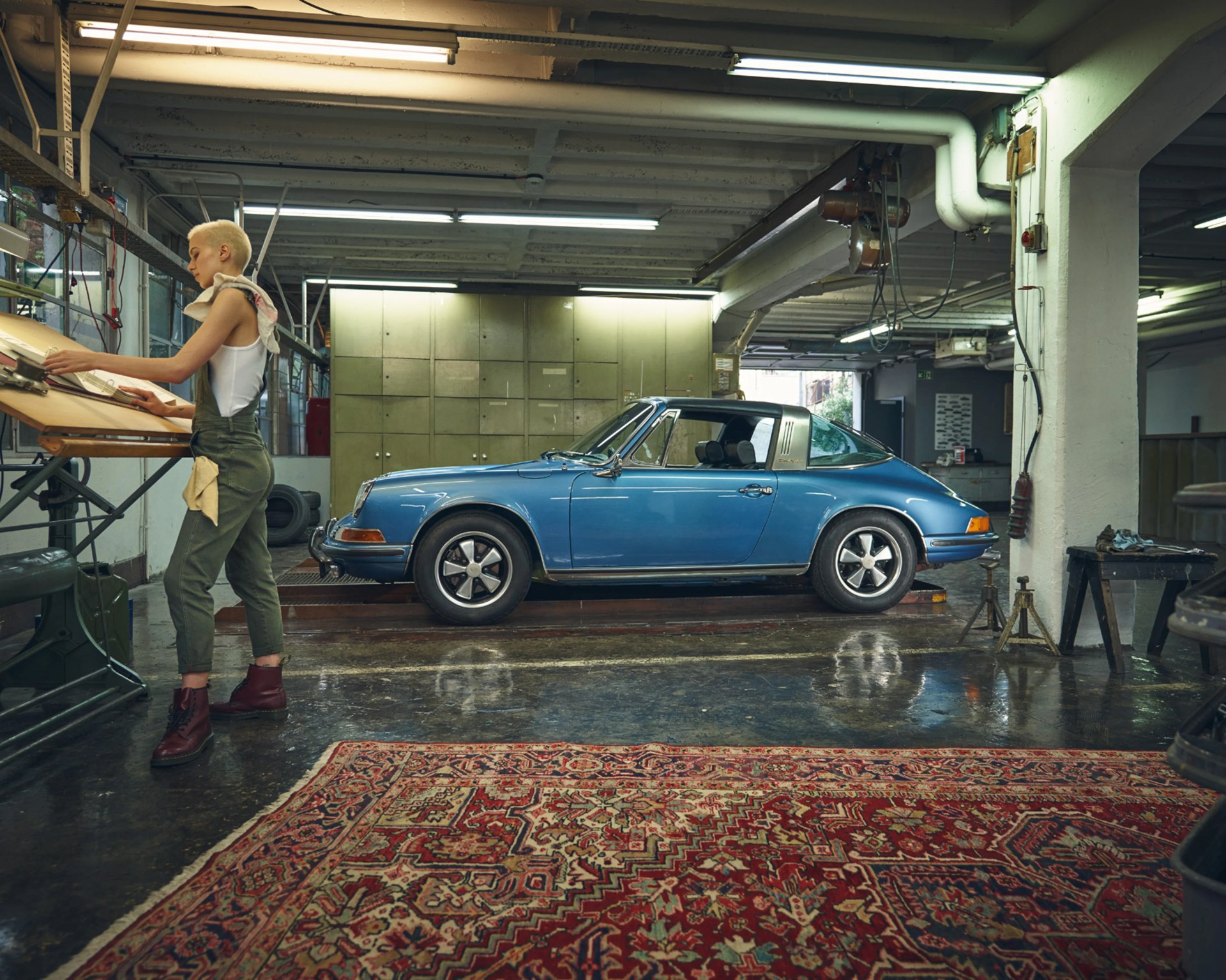 Blue vintage Porsche 911 Targa parked inside a garage with a person working at a drafting table nearby.