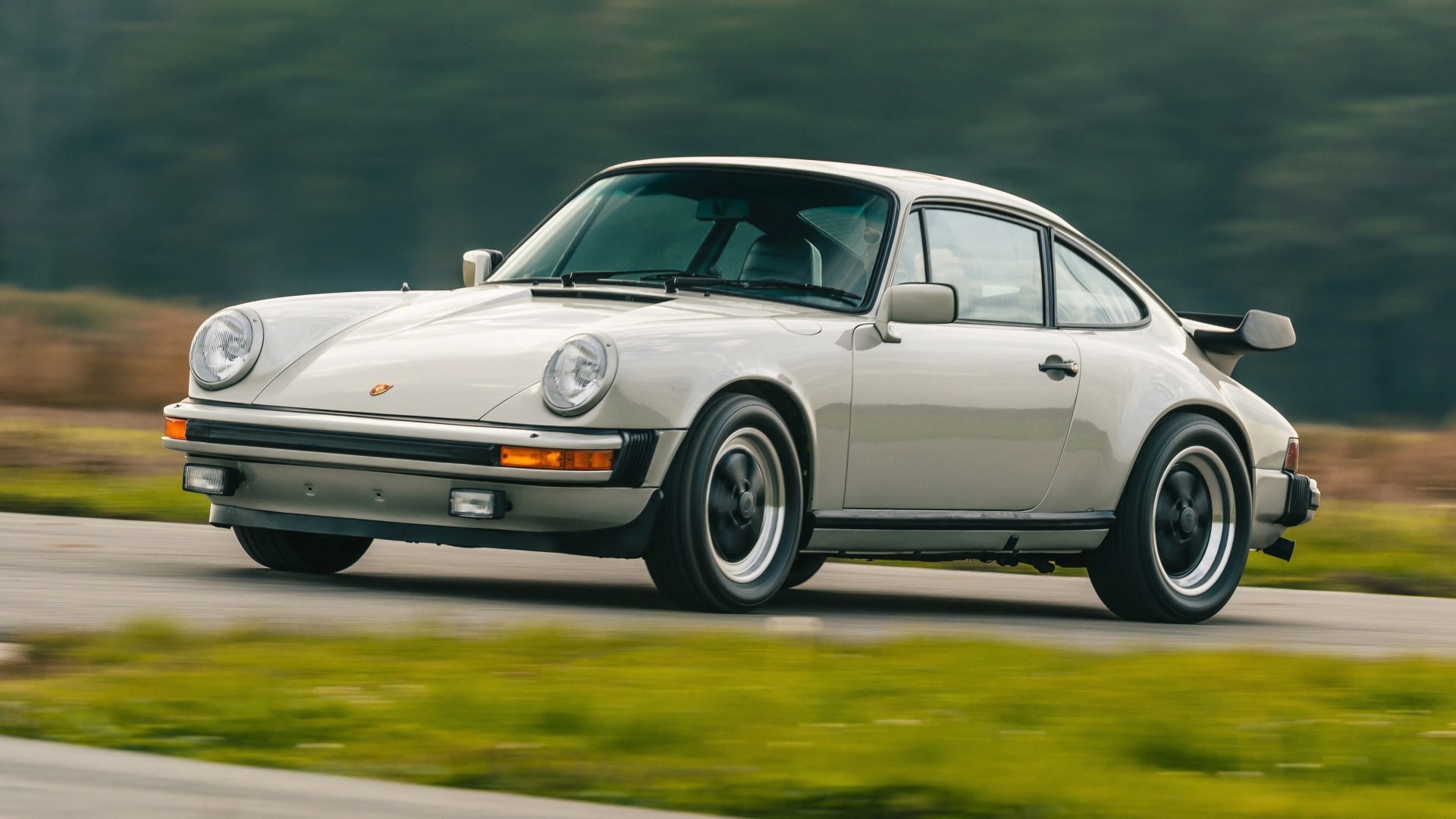 White classic Porsche 911 with black trim and rear spoiler driving on a road with blurred green background.