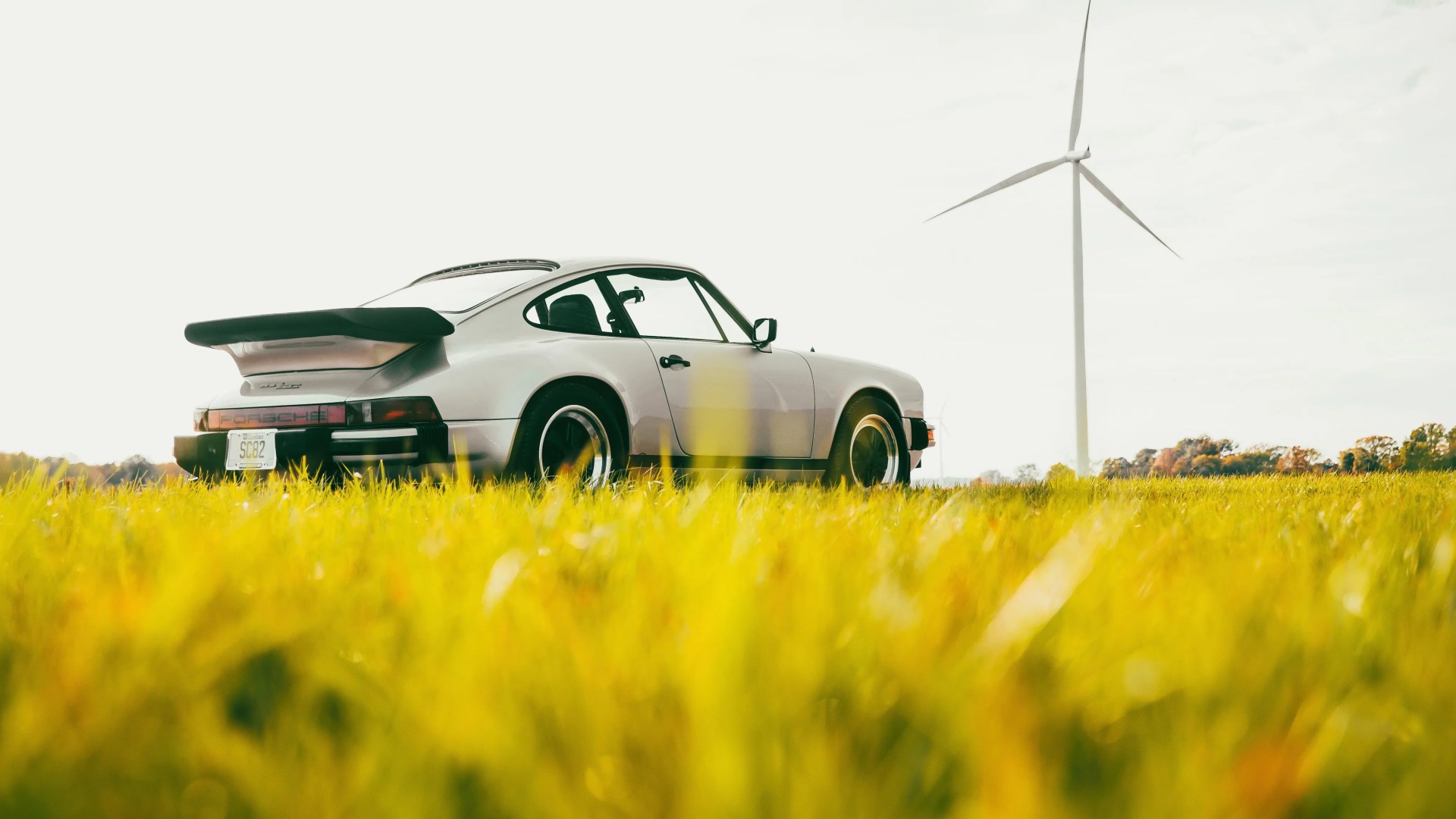 Silver Porsche 911 with black spoiler parked in a yellow grassy field with a wind turbine in the background.