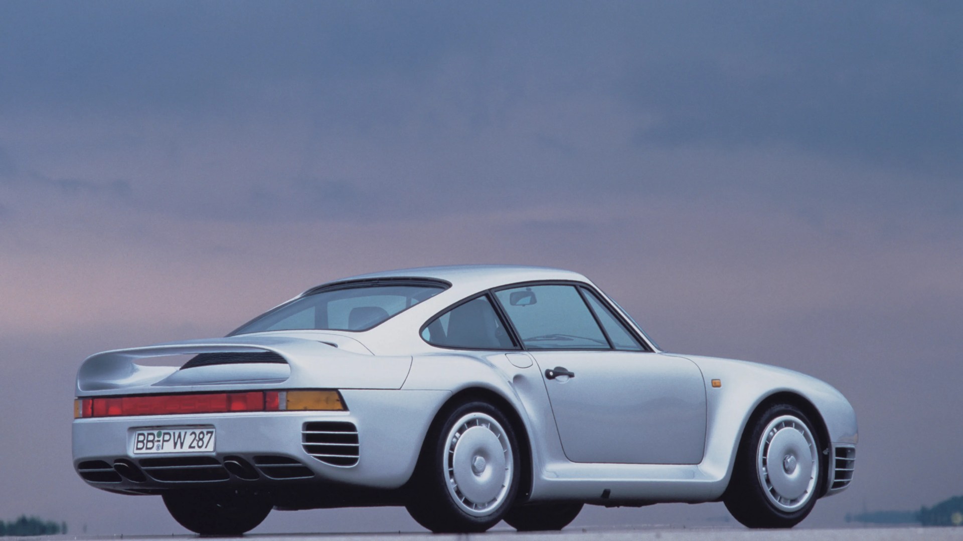 Silver Porsche 959 sports car viewed from the rear side against a dusky sky.