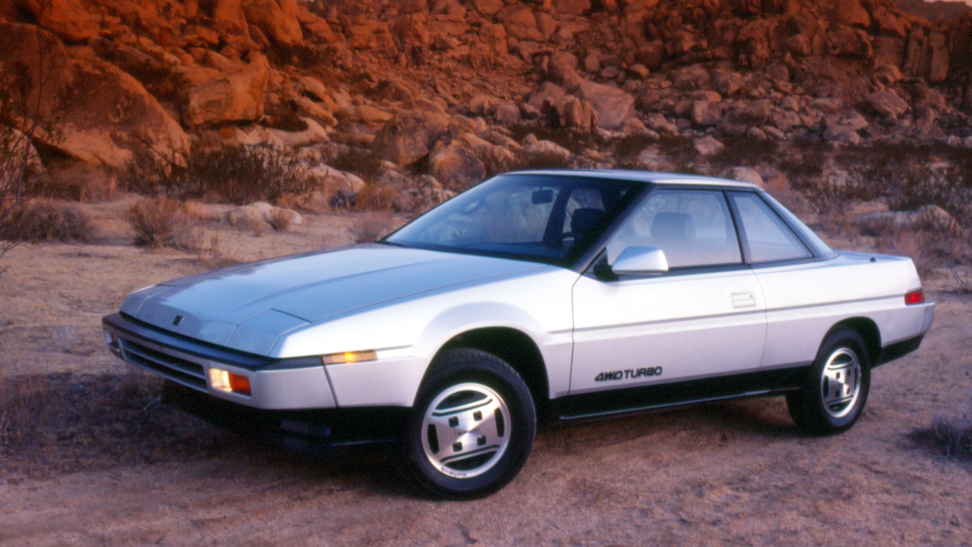 White 1980s two-door coupe car with "4WD Turbo" badge parked on rocky desert terrain.