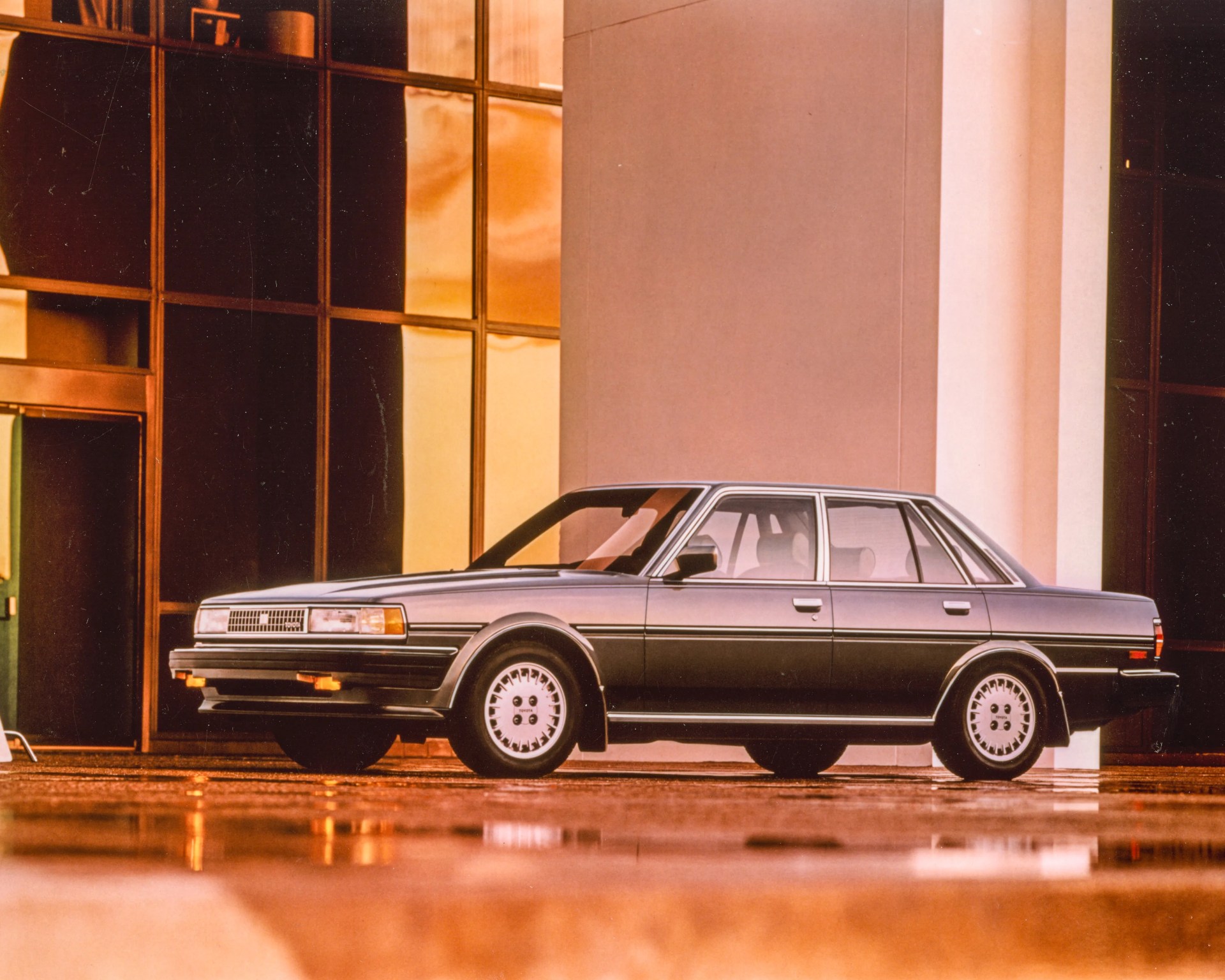 Silver 1980s Toyota sedan parked in front of a building with large reflective windows.