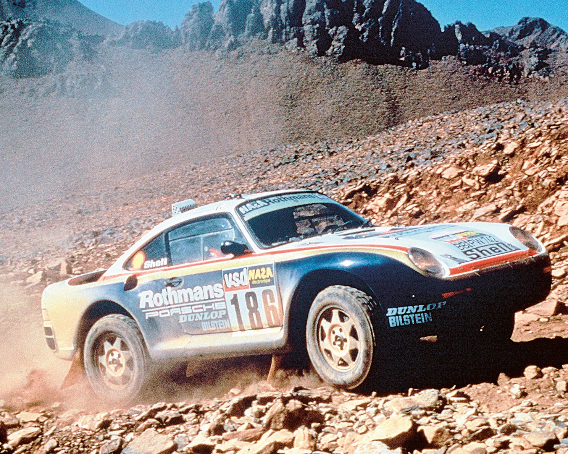 White and blue Rothmans Porsche rally car with number 186 driving on rocky terrain.