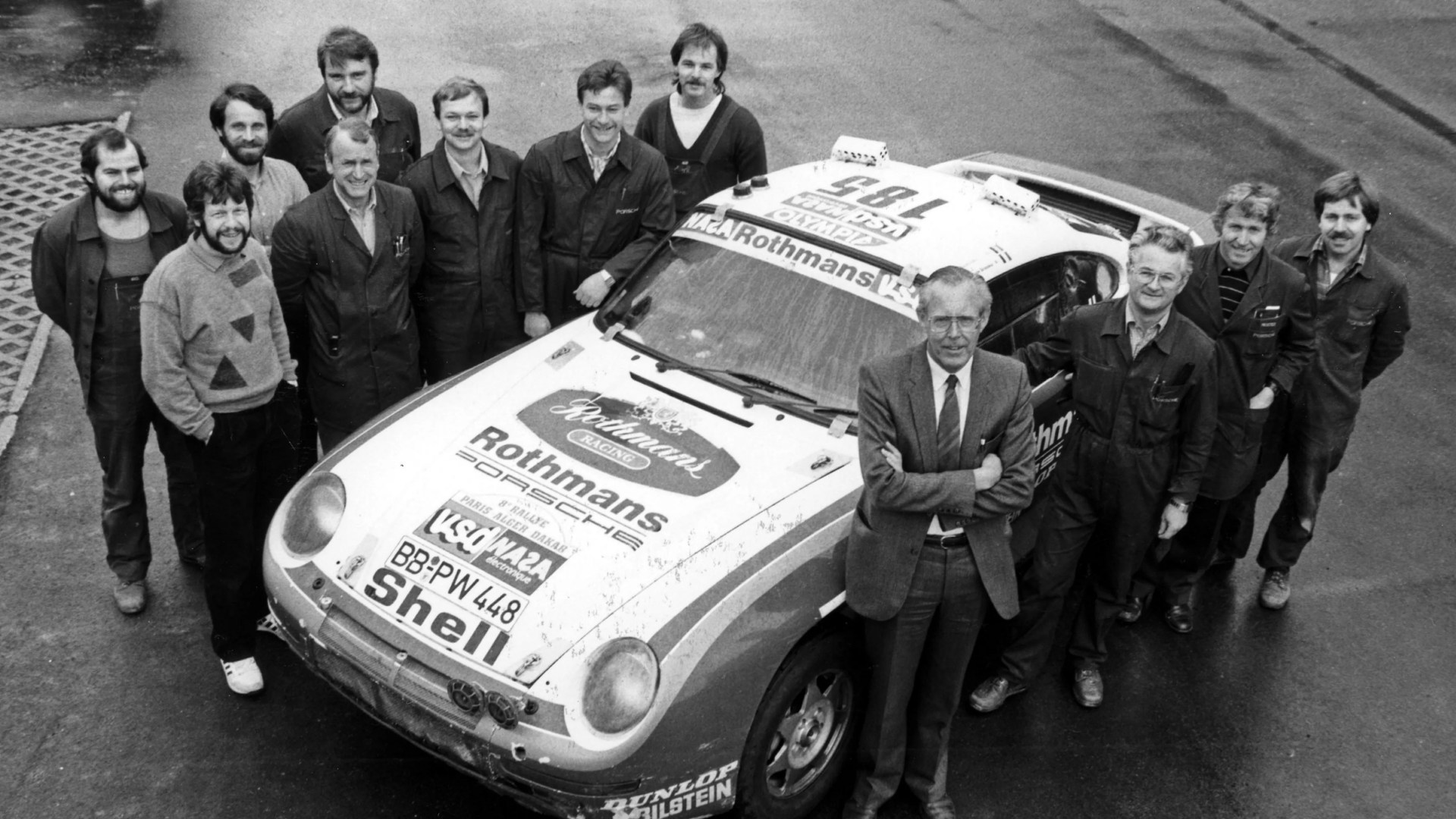 Group of men standing around a vintage Rothmans Porsche race car with racing decals and sponsor logos.