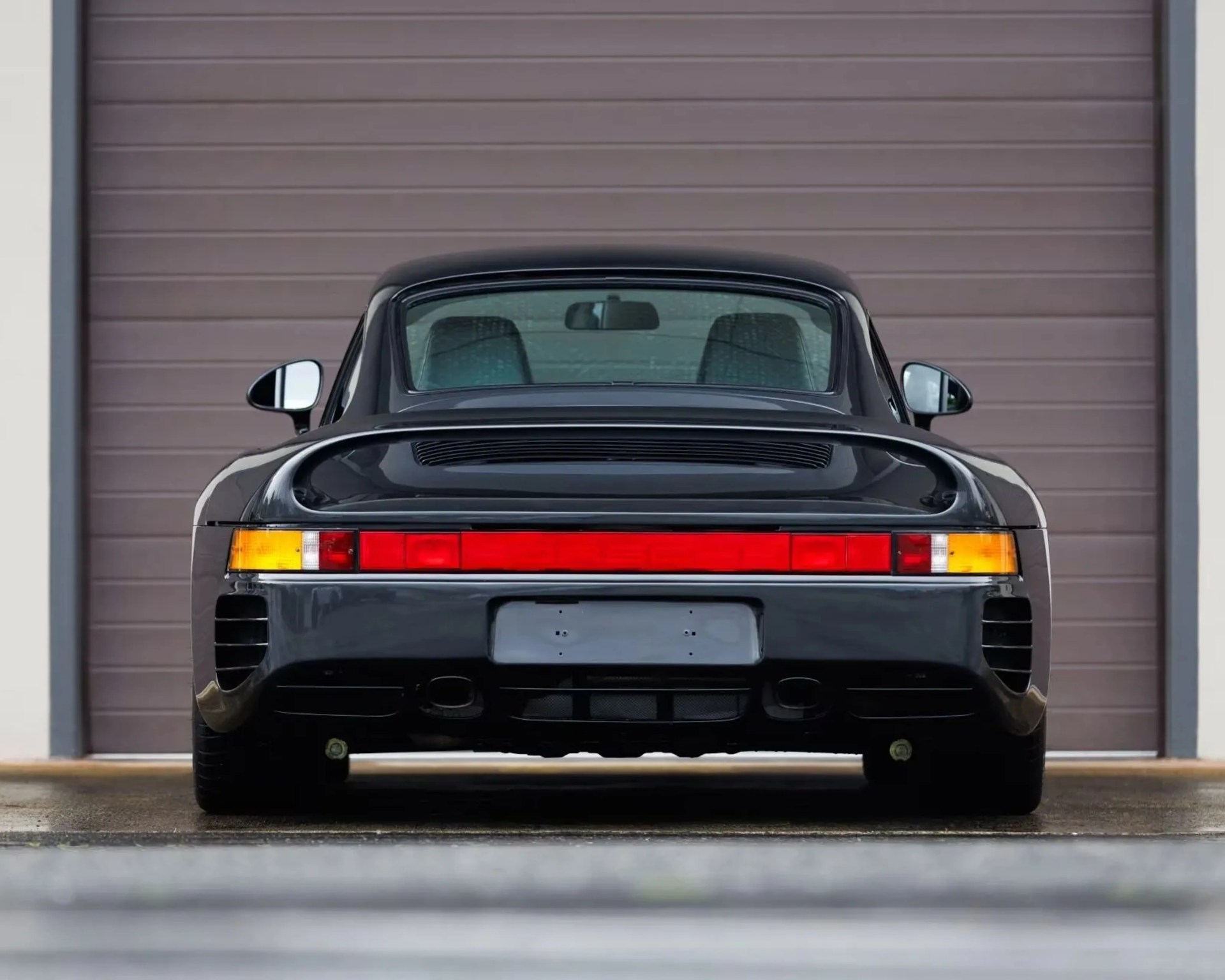 Rear view of a black Porsche 959 sports car with red taillights and dual exhausts in front of a closed garage door.