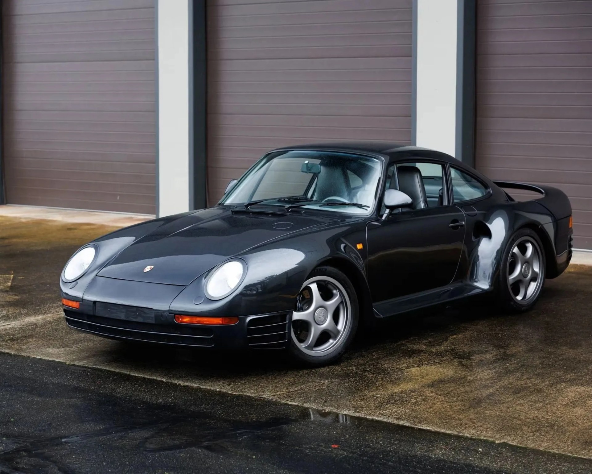 Black Porsche 959 sports car parked on wet pavement in front of closed brown garage doors.