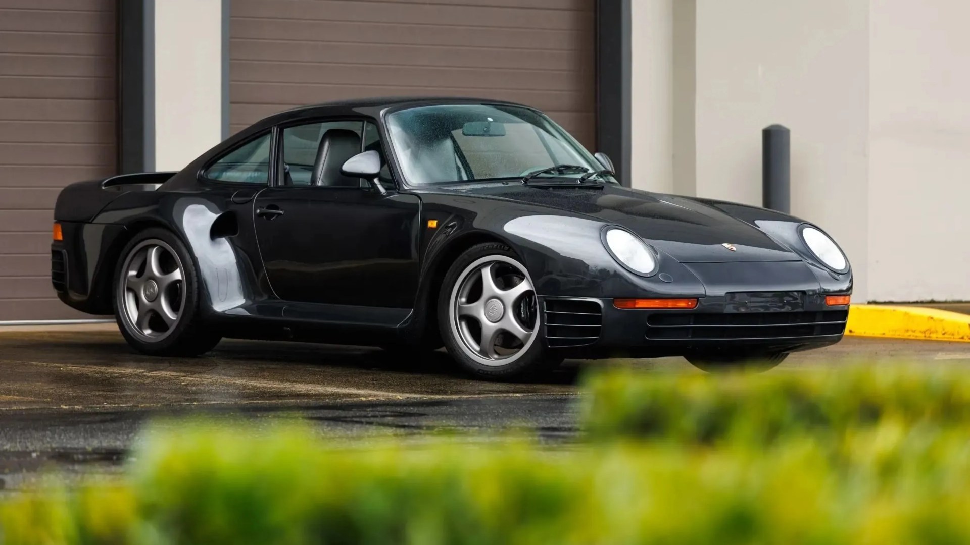 Black Porsche 959 sports car parked on wet pavement in front of a brown garage door.