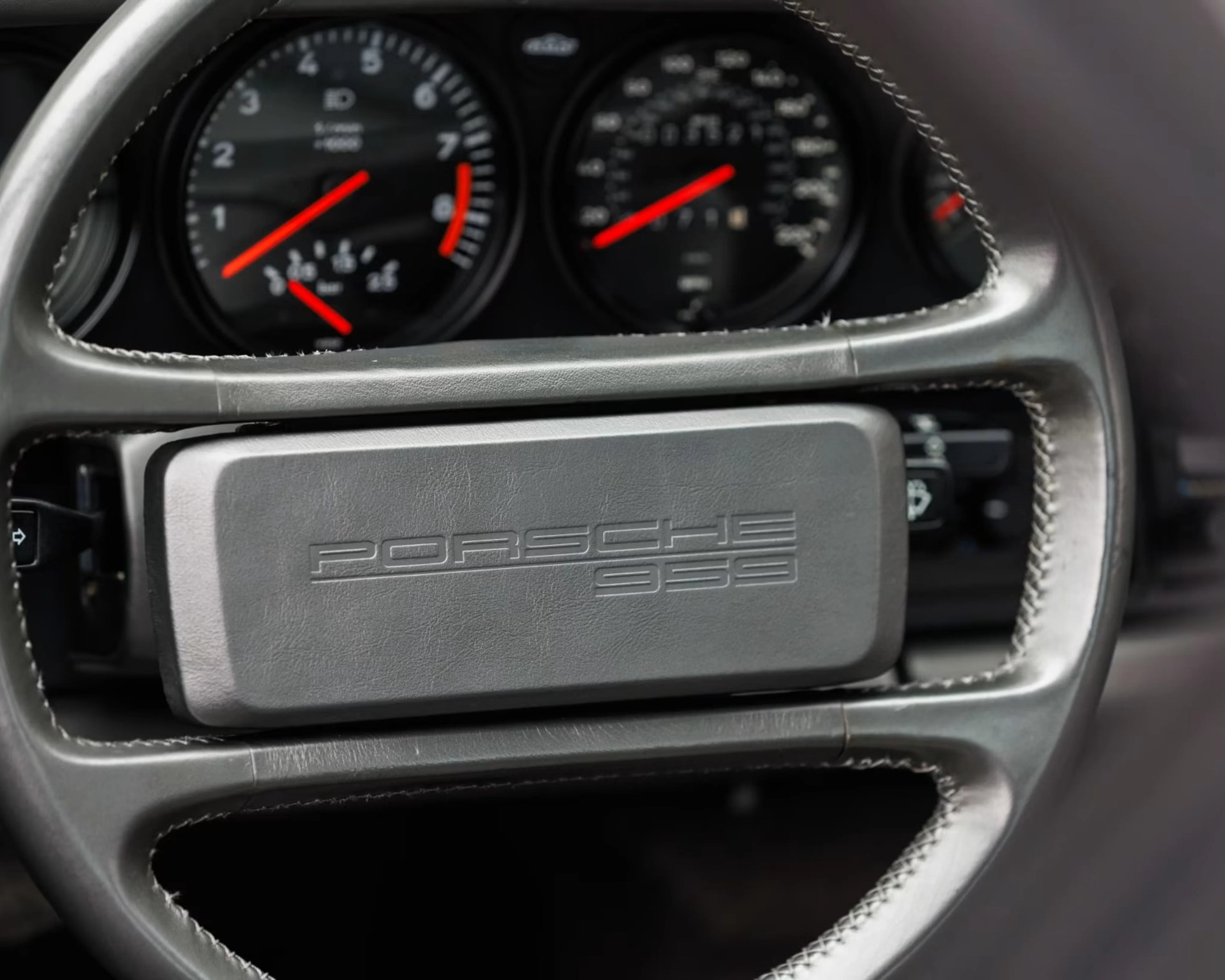 Gray leather steering wheel with "Porsche 959" embossed on the center airbag cover and red gauge needles in the background.